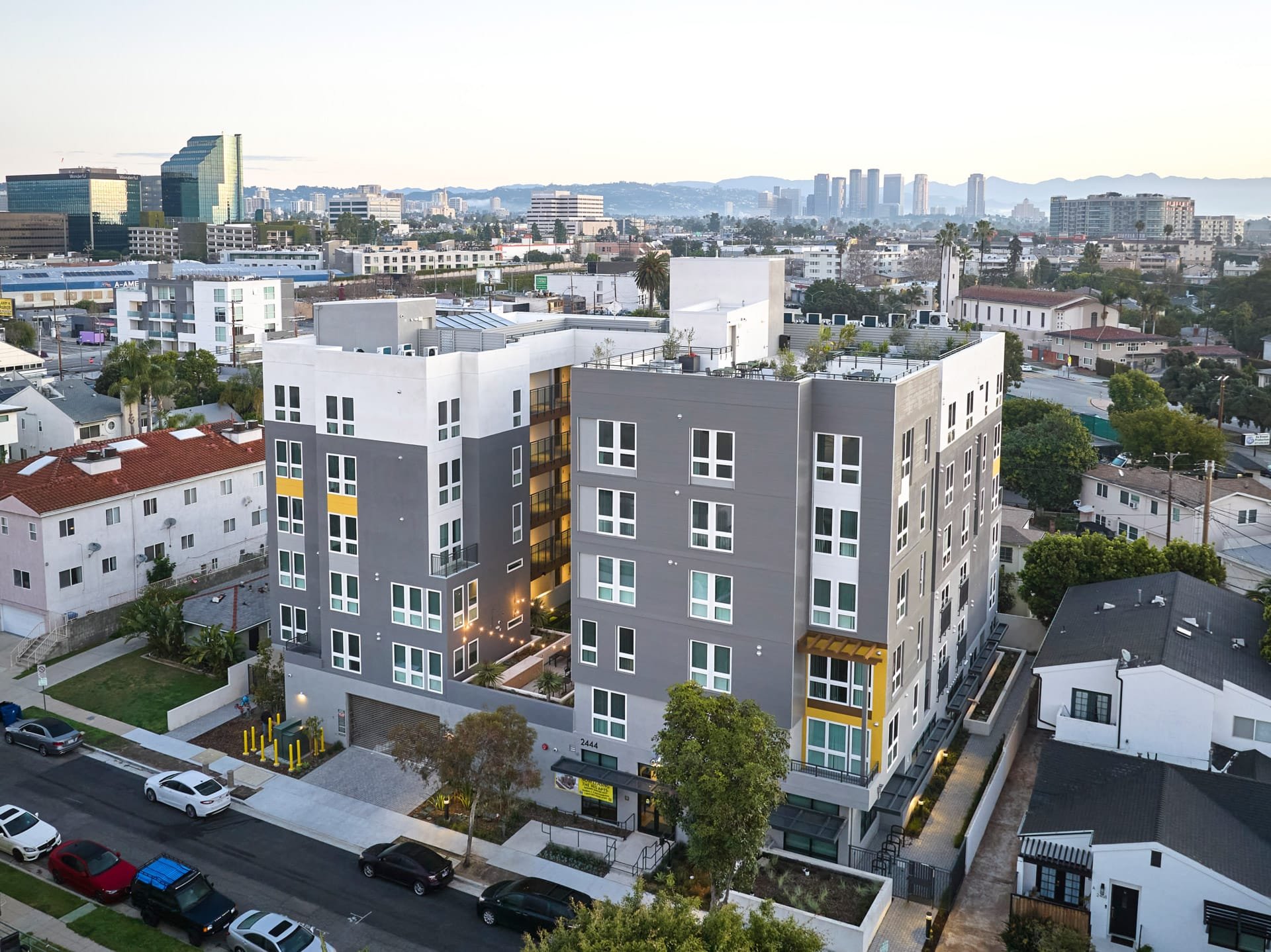 A modern multi-story apartment building with gray and white exterior, yellow accents, and a rooftop garden, situated in an urban neighborhood with cars parked on the street and other residential structures nearby.
