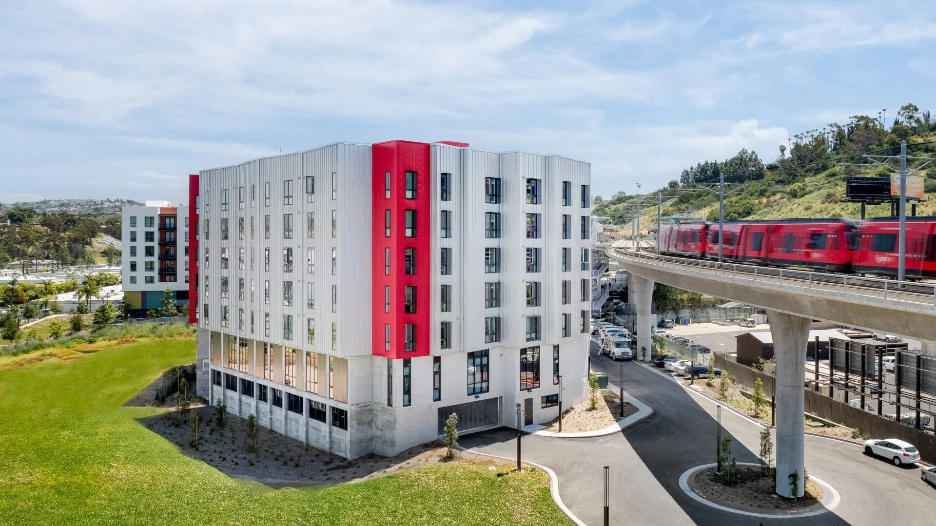 Modern multi-story apartment building with a curved design, mostly white with red accents, situated next to an elevated train track with a red train passing by, surrounded by greenery and a parking lot.