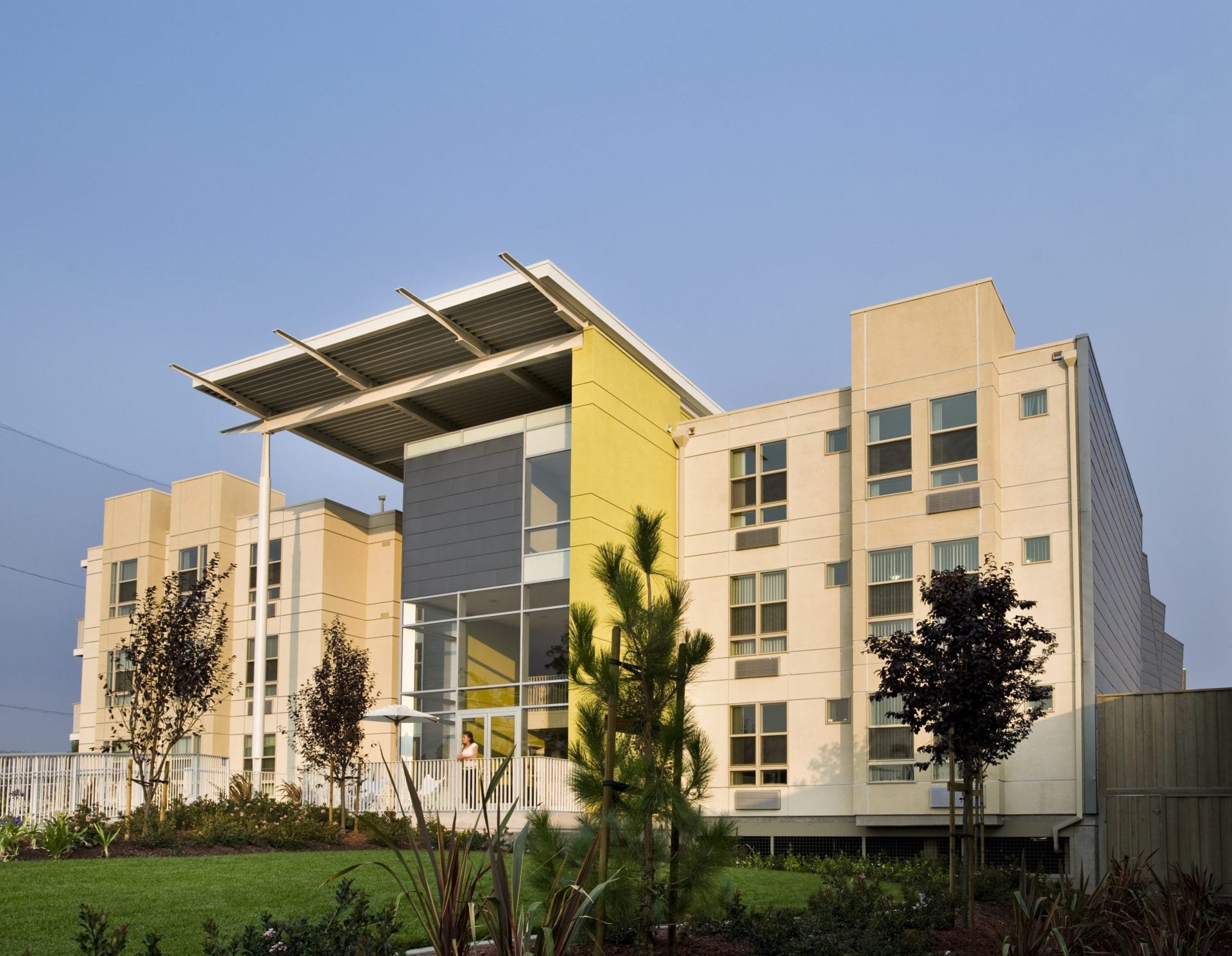 Modern multi-story apartment building with a yellow accent wall and a large overhanging roof, surrounded by landscaped trees and a lawn, under a clear blue sky.