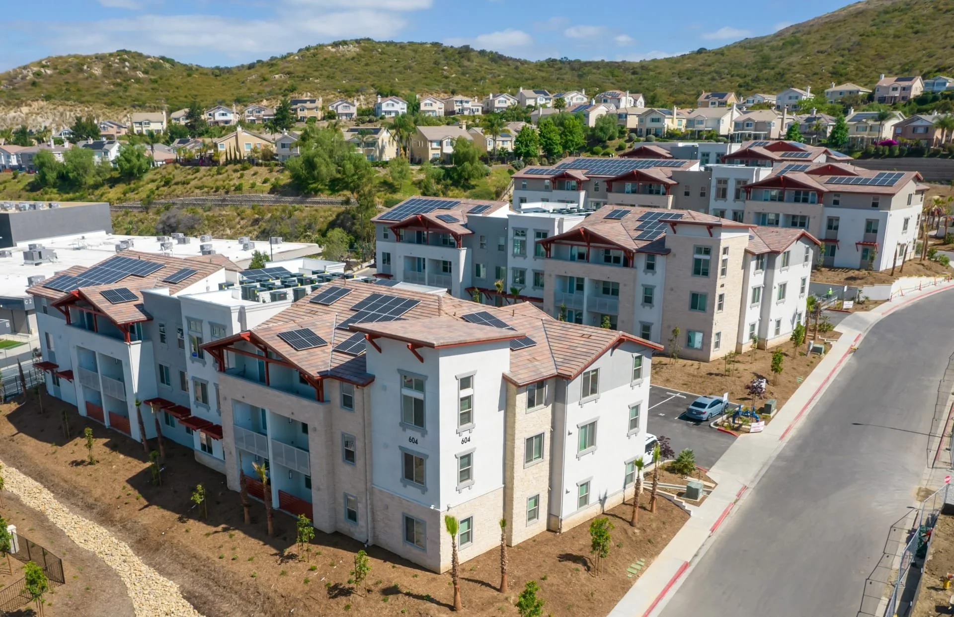 Aerial view of newly constructed apartment buildings with solar panel rooftops, surrounded by small trees and parked cars, in a hilly residential area.