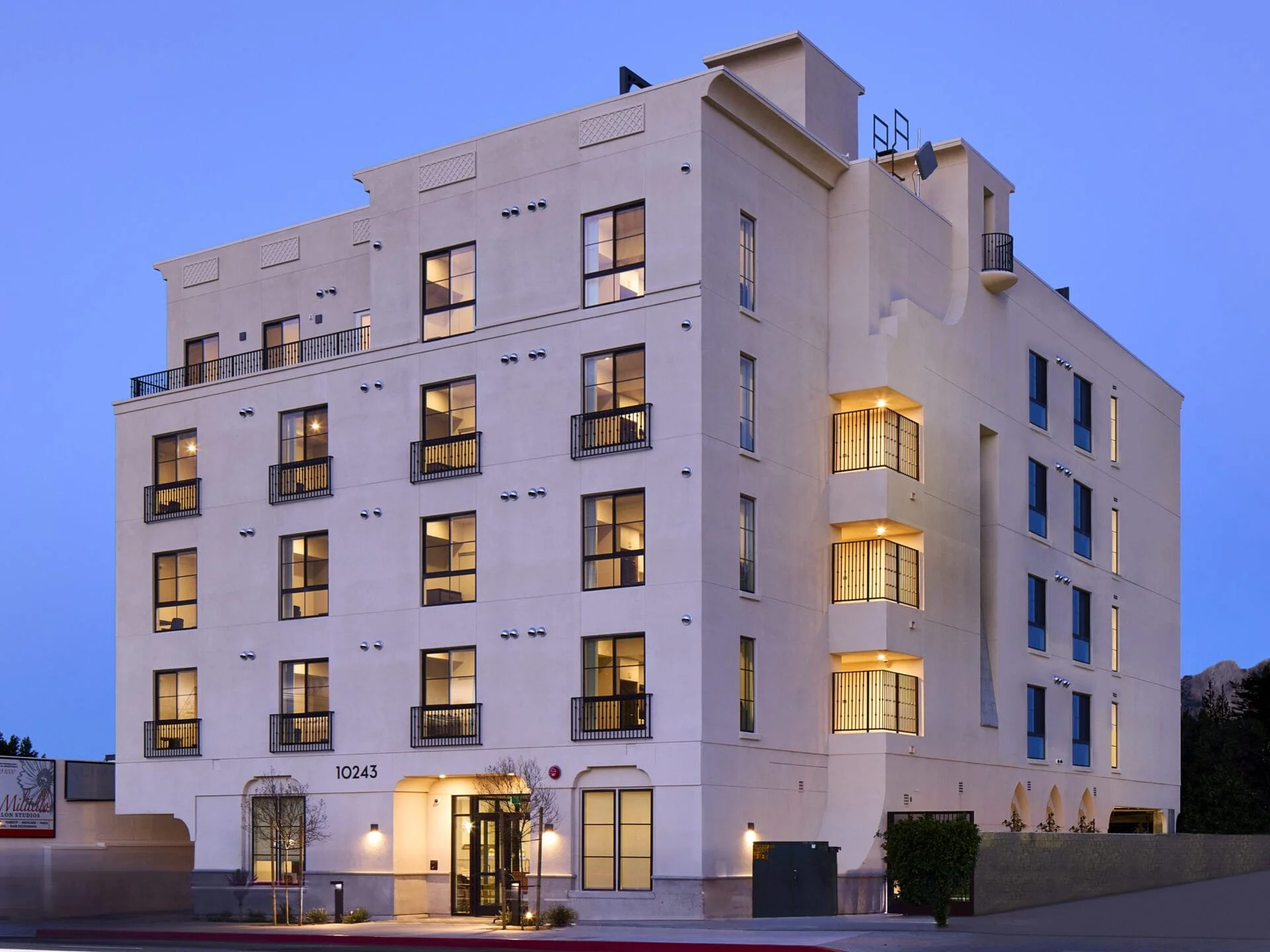 A modern multi-story apartment building with lit windows and balconies, located on a street during dusk with a clear blue sky.