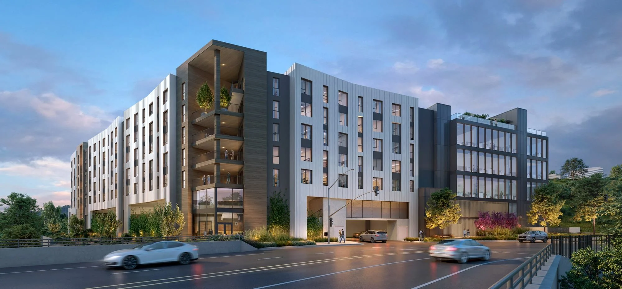 Modern multi-story apartment building with glass windows, balconies, and greenery, situated beside a busy street with cars and pedestrians, during dusk.