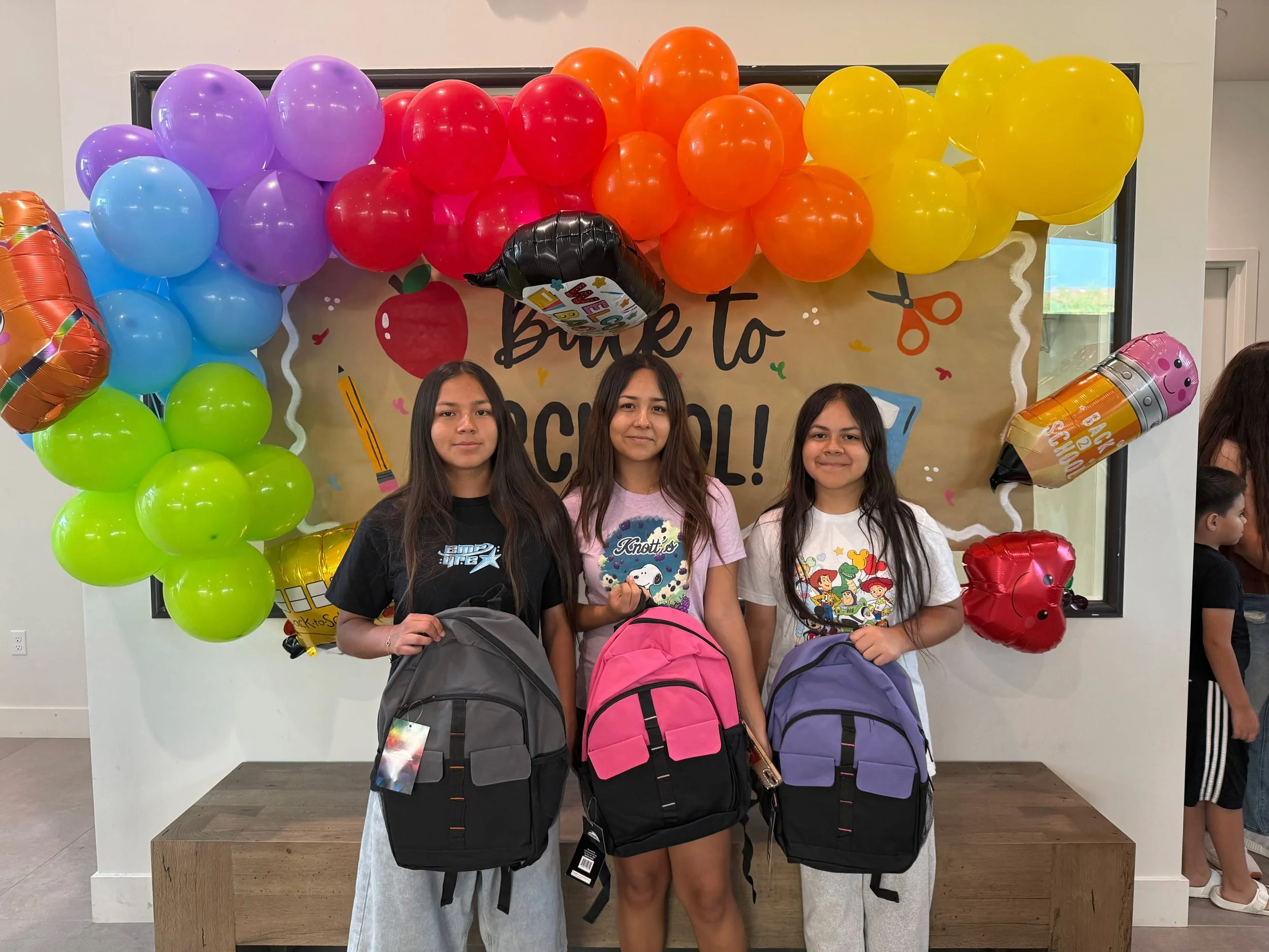 Three girls stand in front of a colorful school-themed balloon display holding backpacks for a back-to-school event.