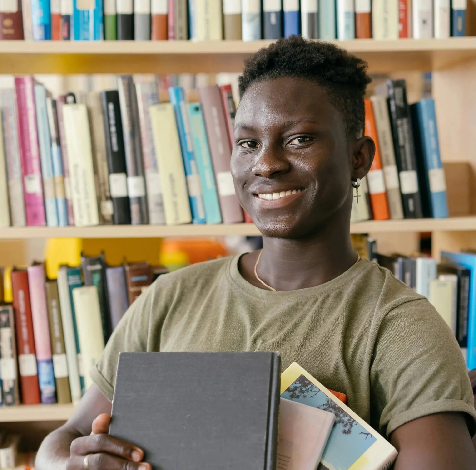 A teen holding books in a library, smiling at the camera with bookshelves in the background.