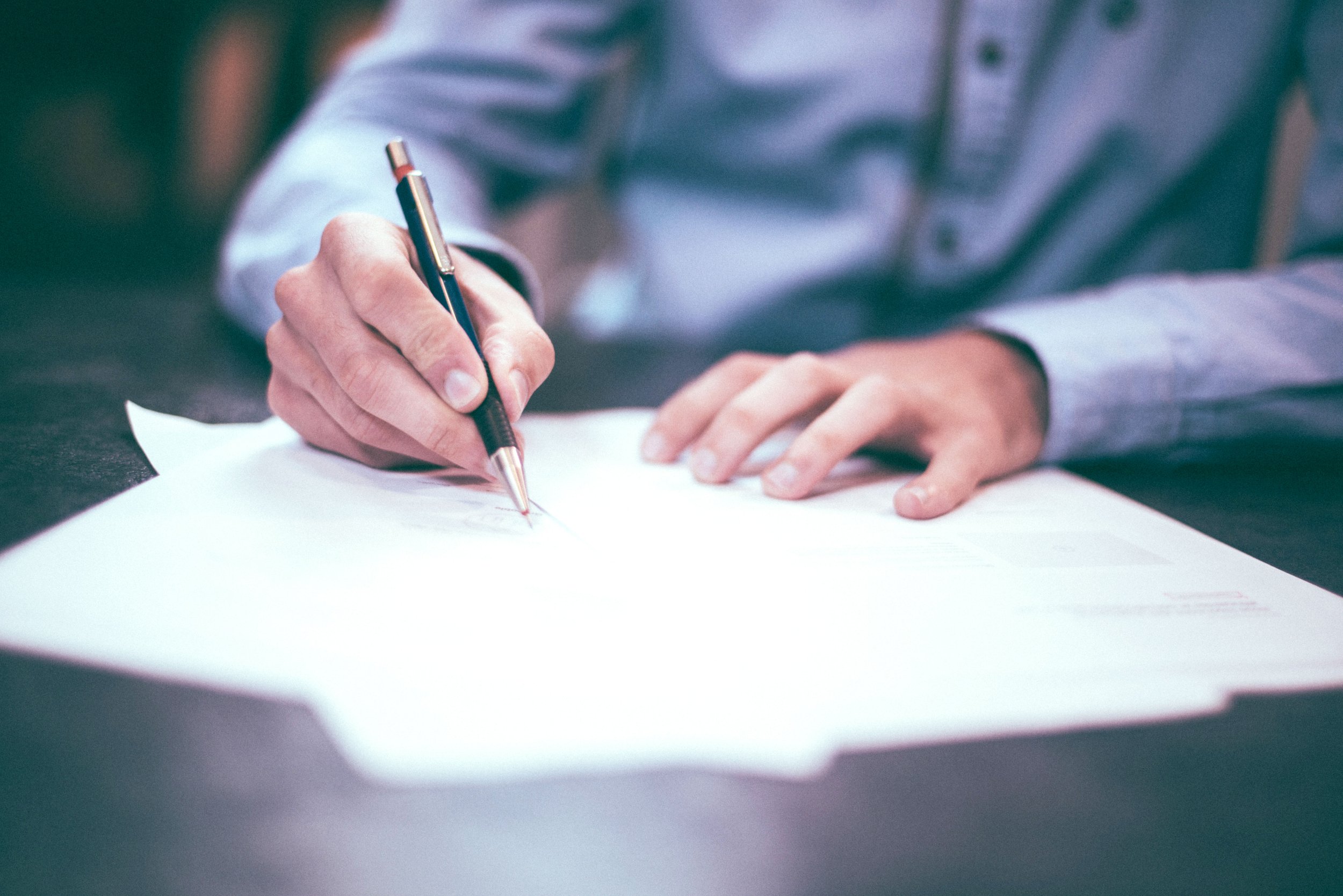 A person in a long-sleeved, button-up shirt writing on a stack of white papers with a black and gold ballpoint pen.