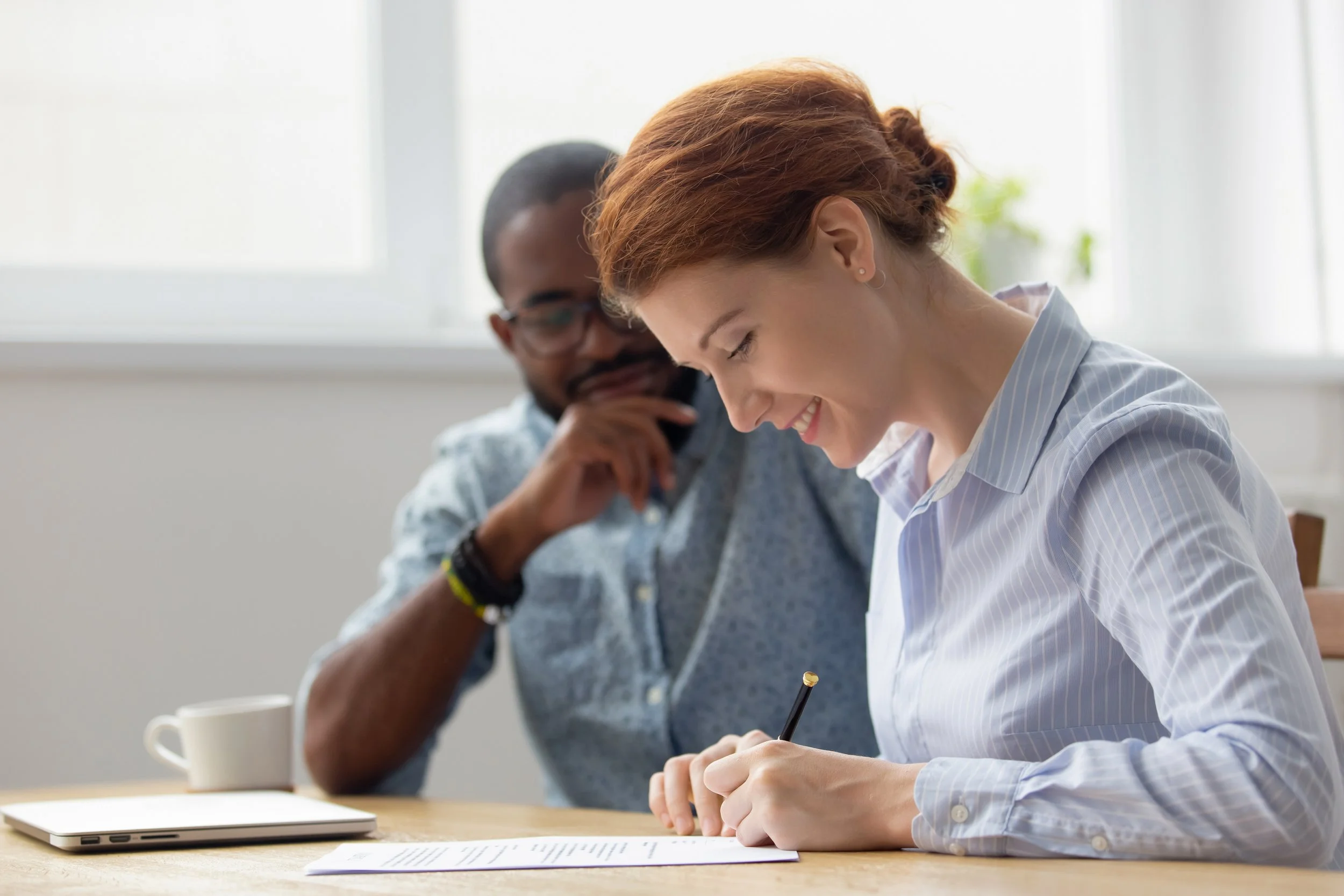 Two colleagues working at a desk, one woman smiling and signing a document, a man observing in the background.