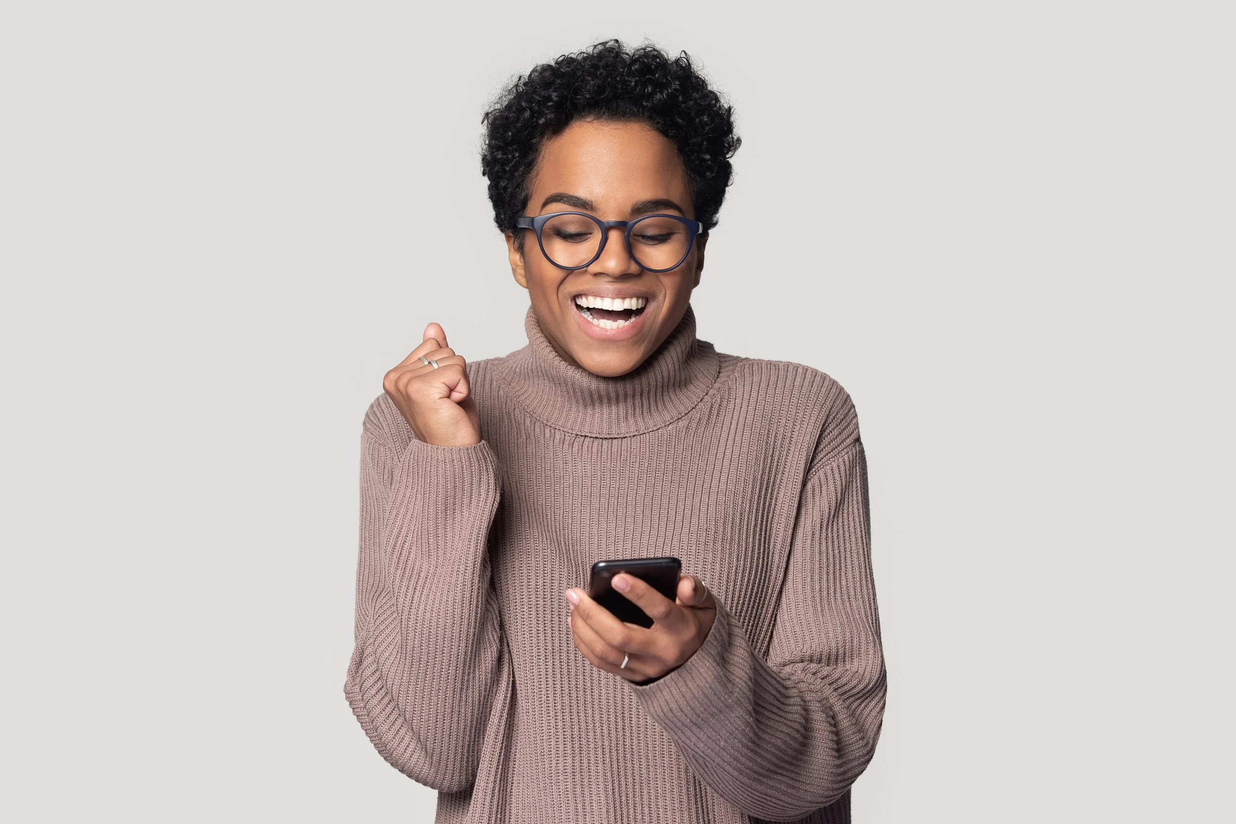A woman with short curly black hair, glasses, and a beige turtleneck sweater looking at her phone, smiling and celebrating.