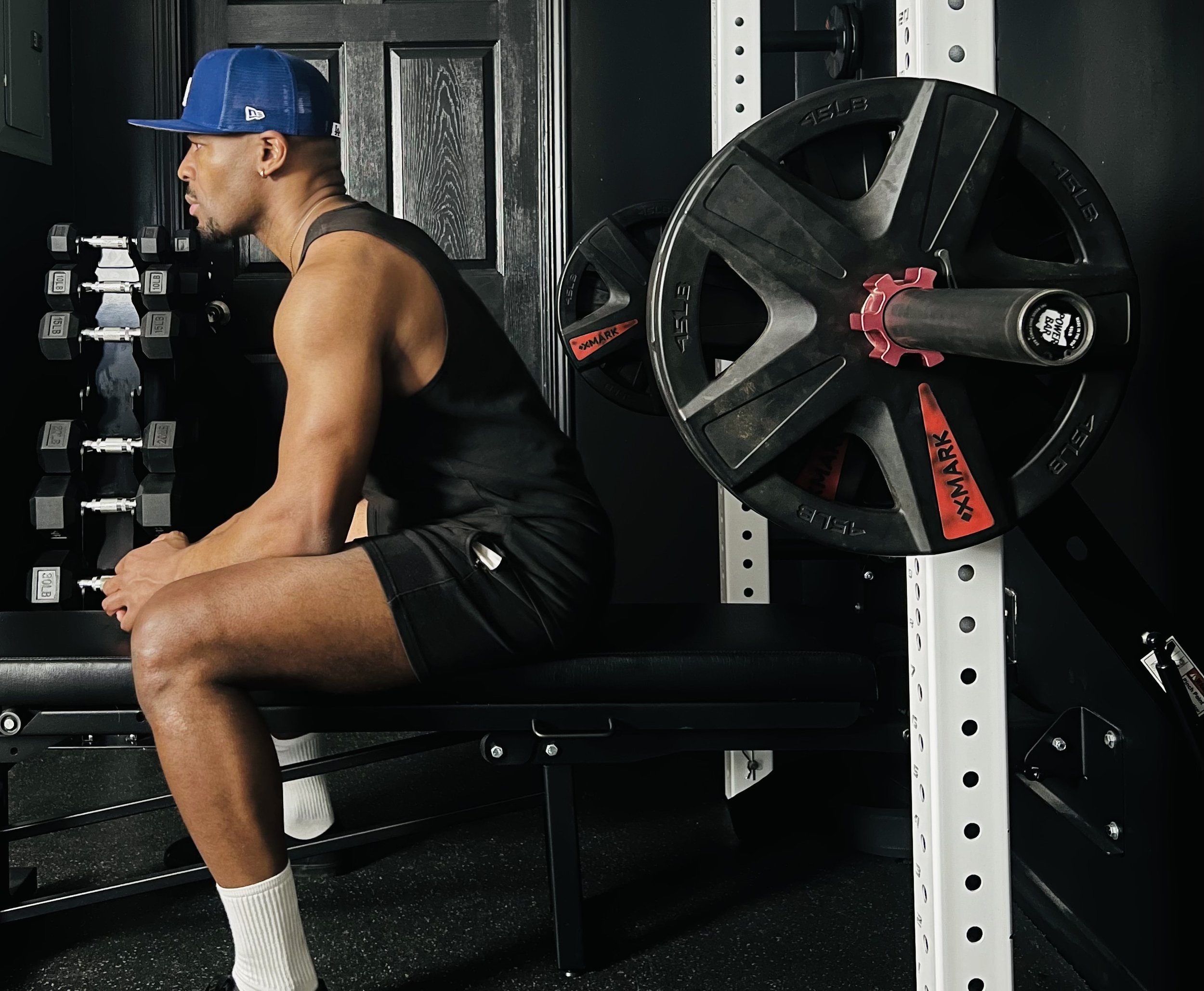 Man sitting on a workout bench in a gym, wearing a blue cap, black tank top, black shorts, and white socks, with gym equipment including dumbbells and weight plates around him.