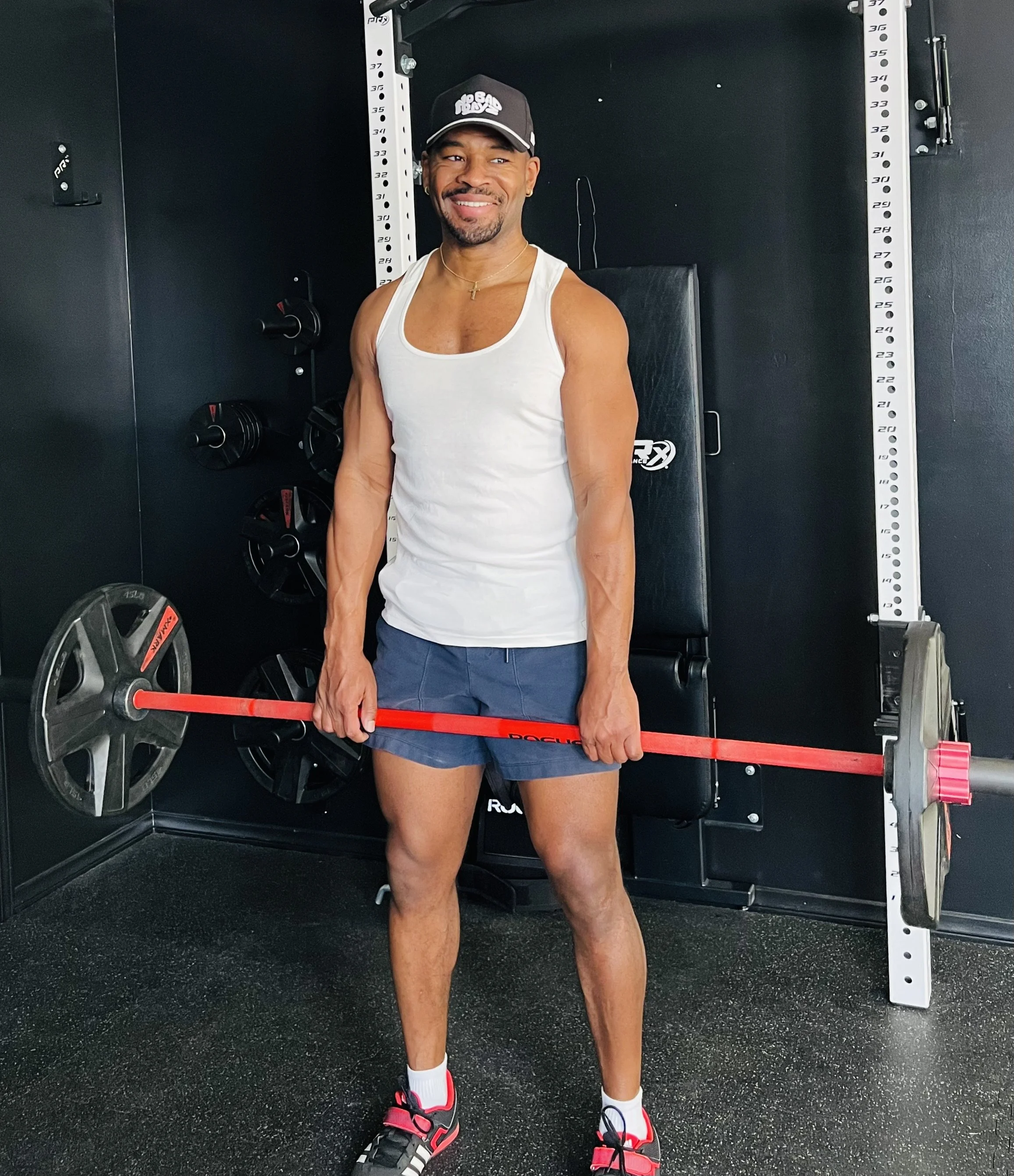 Man in a white tank top and navy shorts holding a barbell inside a gym with black walls and workout equipment.