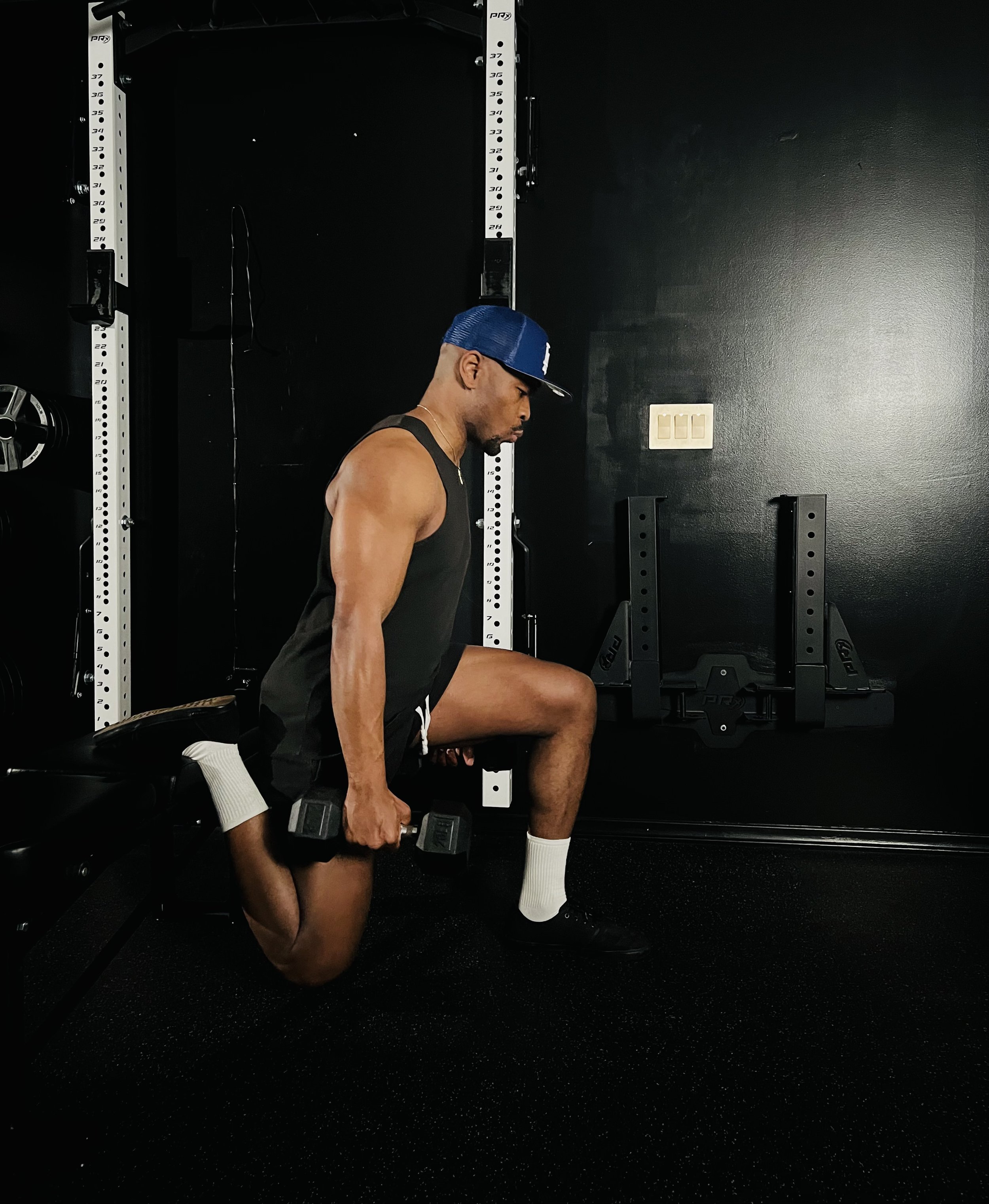 A man performing a split squat exercise with dumbbells in a home gym with black walls and equipment.