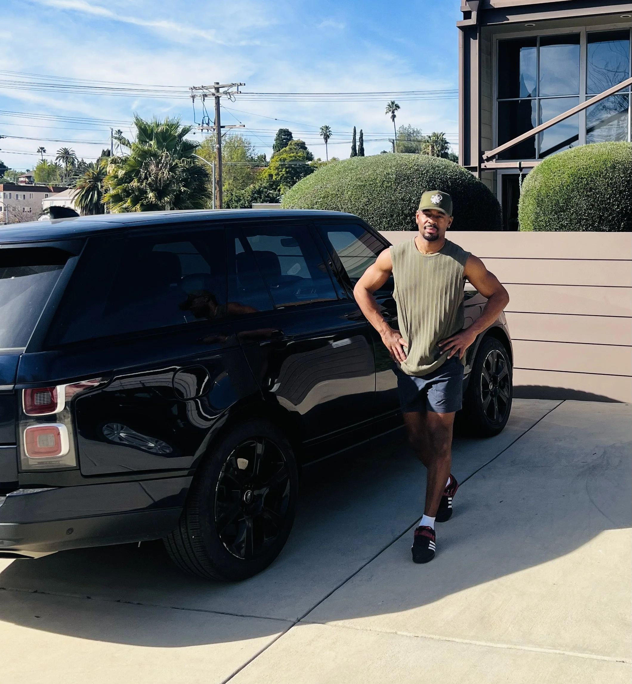 A man standing next to a black luxury car outdoors on a sunny day with a residential building, green bushes, and palm trees in the background.