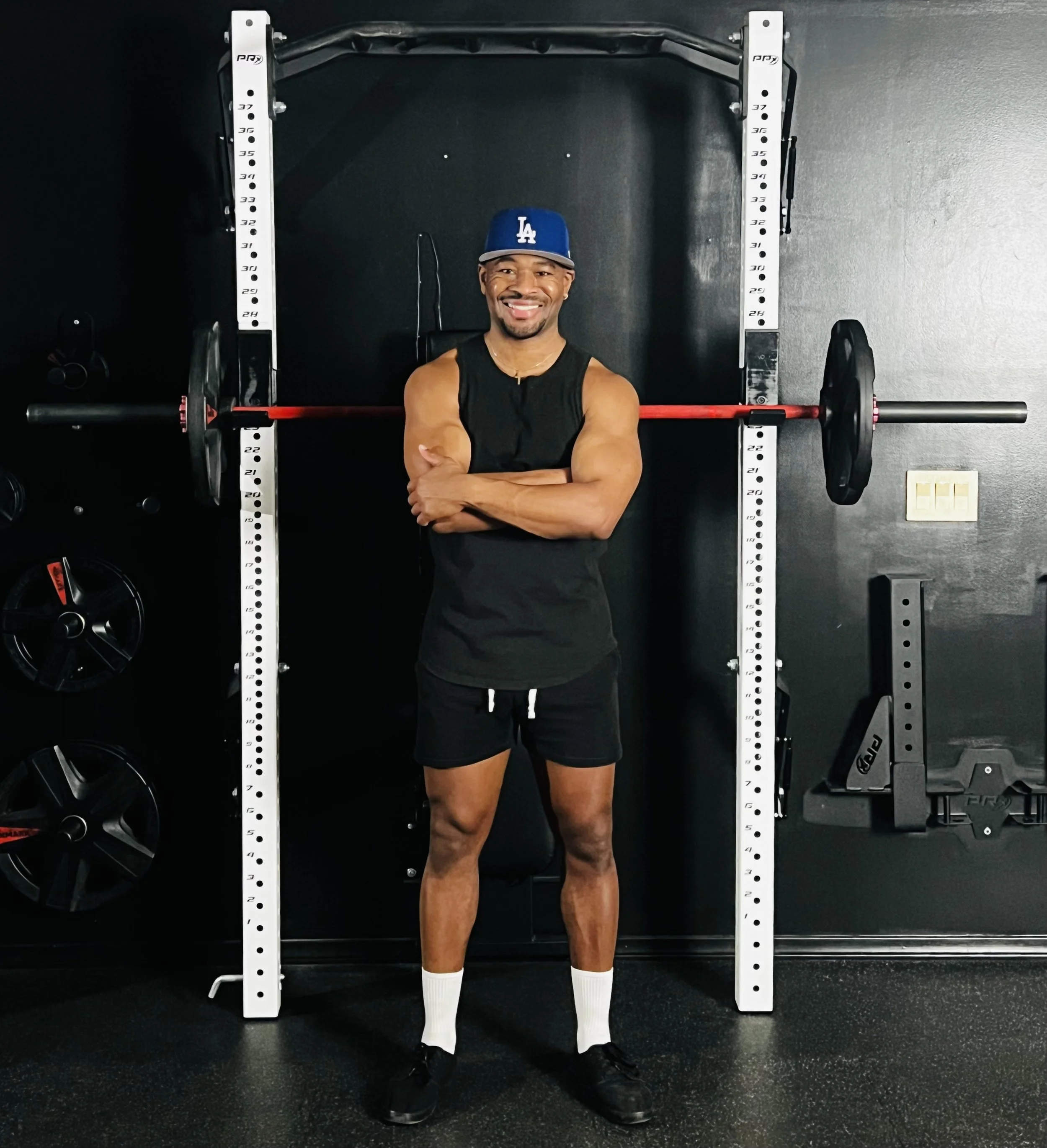 Man in athletic wear, standing in front of a power rack with a barbell, smiling with arms crossed in a gym.
