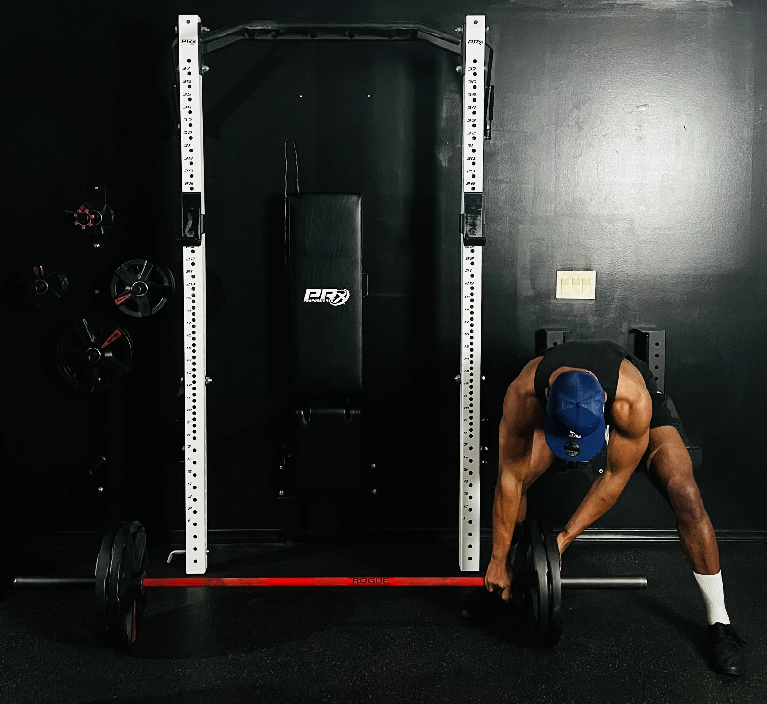 A man bending over, preparing to lift weights in a gym with black walls, a power rack, weight plates, a bench, and a red barbell on the floor.