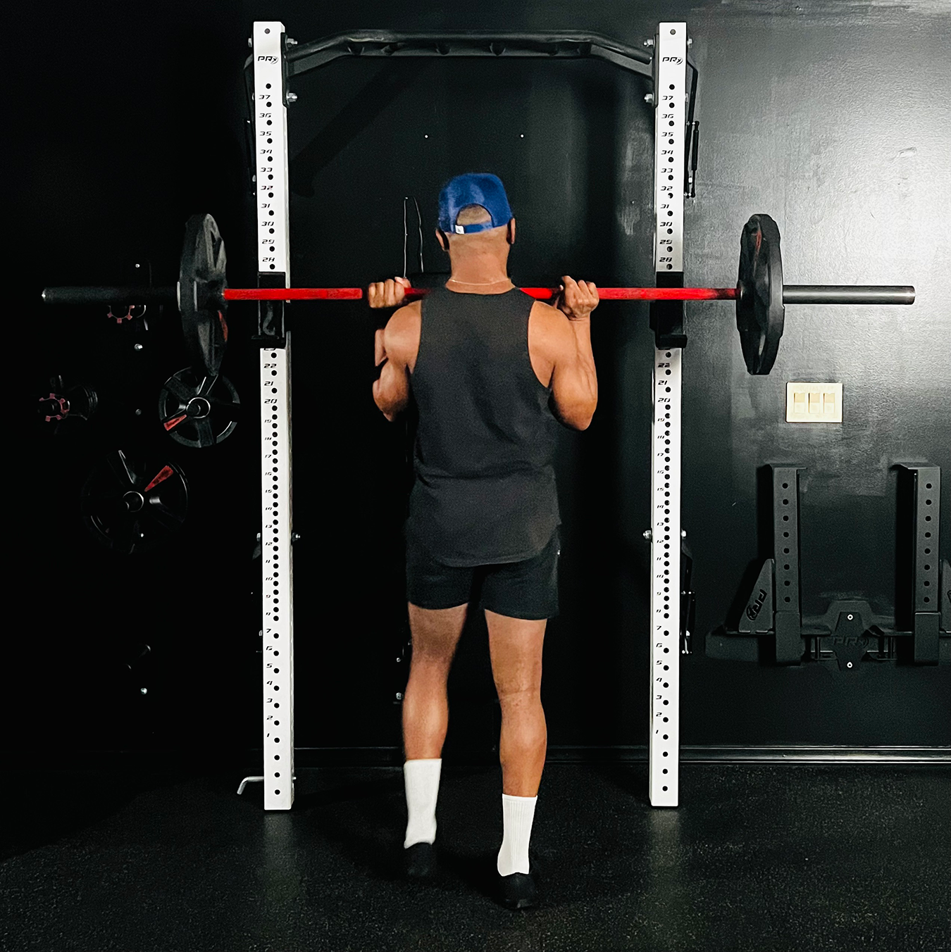 A man lifting a barbell with weights at a gym, facing away from the camera, wearing a black tank top, black shorts, white socks, black shoes, and a blue cap.