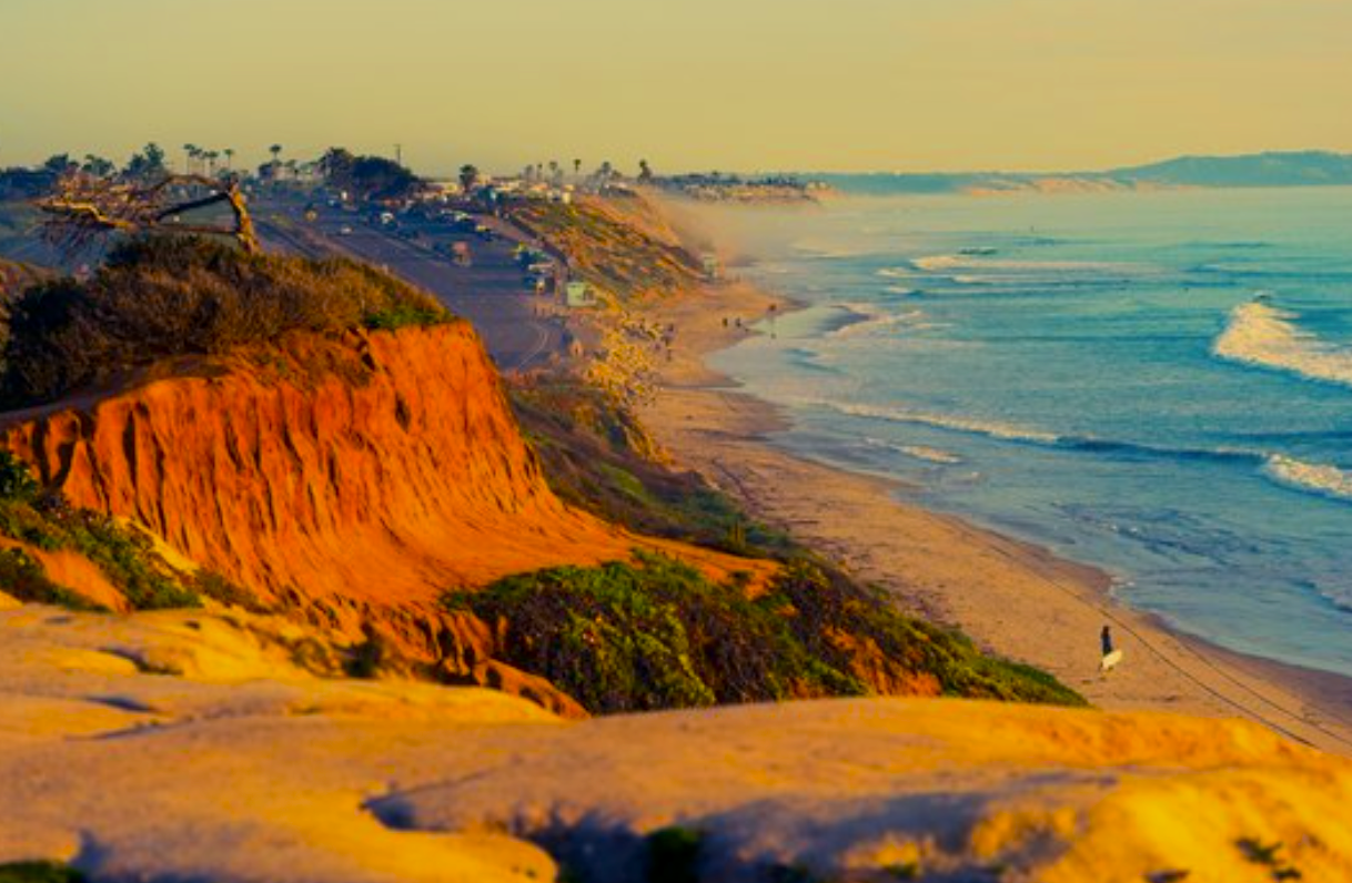 Sunset on the California coastline with orange cliffs, sandy beach, waves, and a person walking a dog.