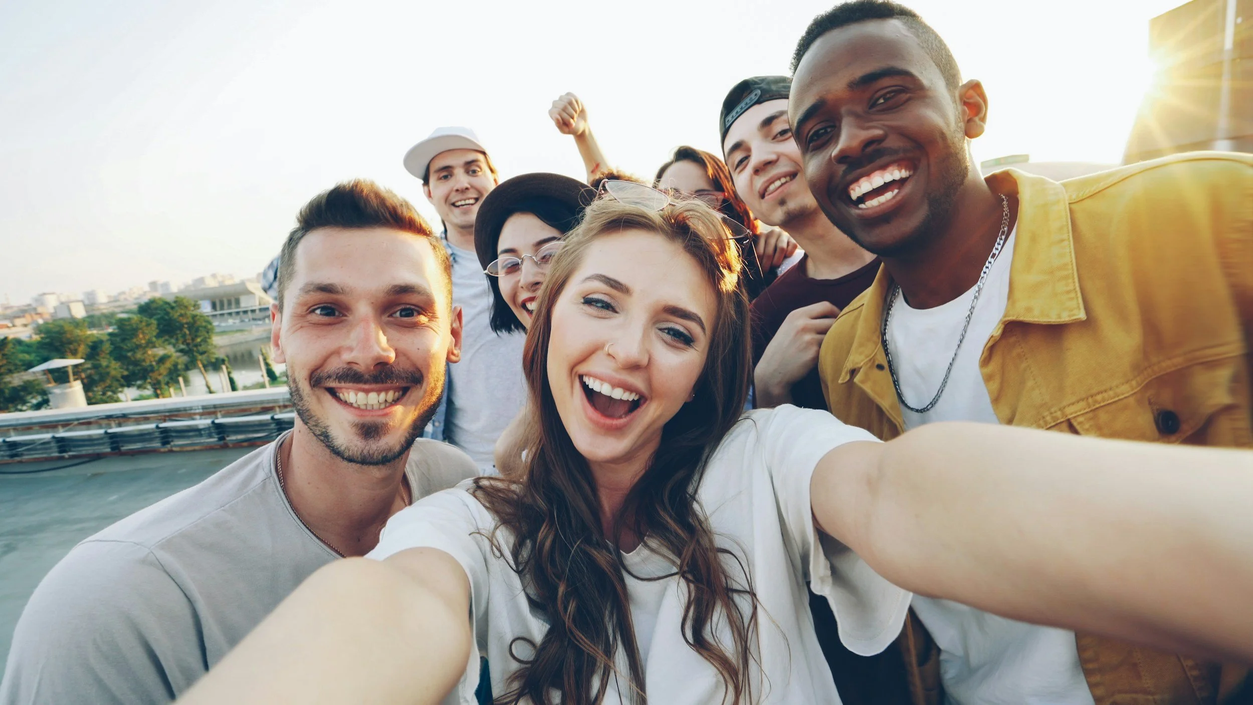 A group of happy young people taking a selfie outdoors during the daytime, with city buildings and trees in the background.