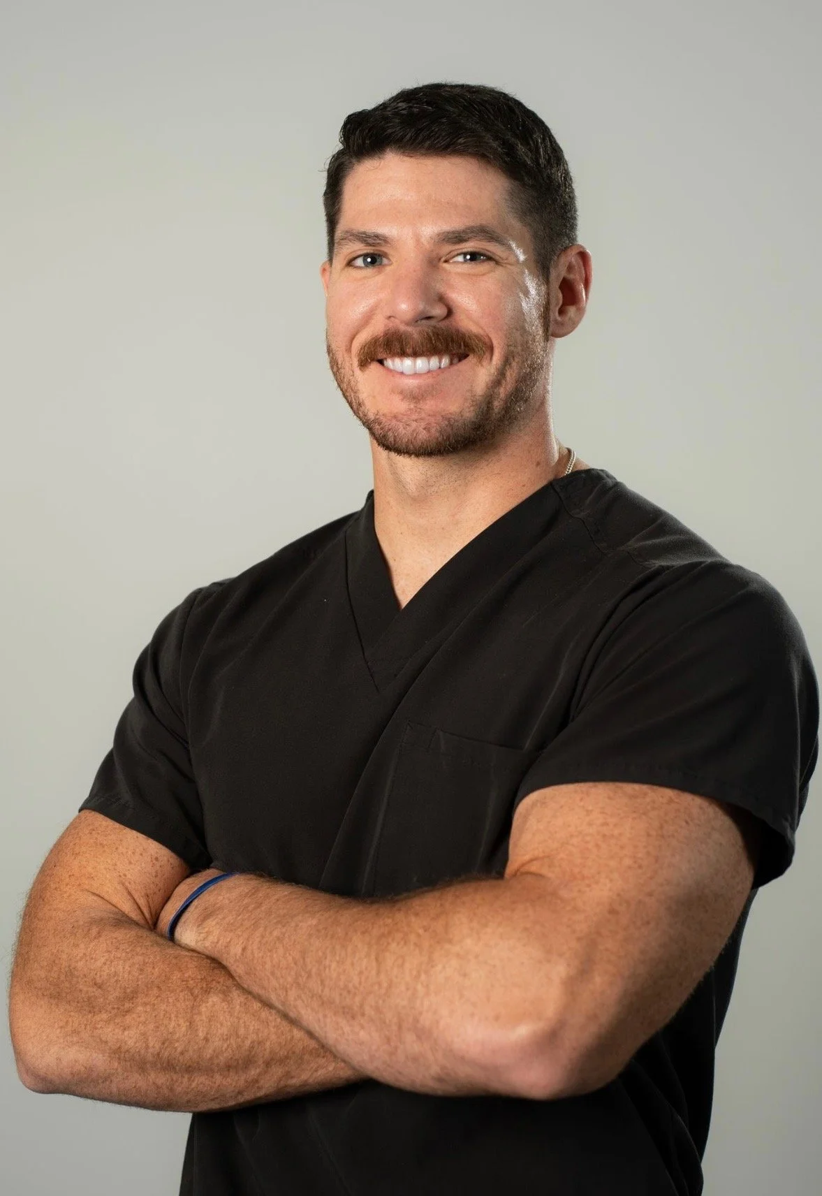 A man with dark hair, a beard, and mustache wearing black medical scrubs, smiling with arms crossed against a plain background.