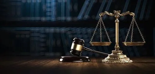 Justice scale and gavel on wooden table with bookshelf in background.