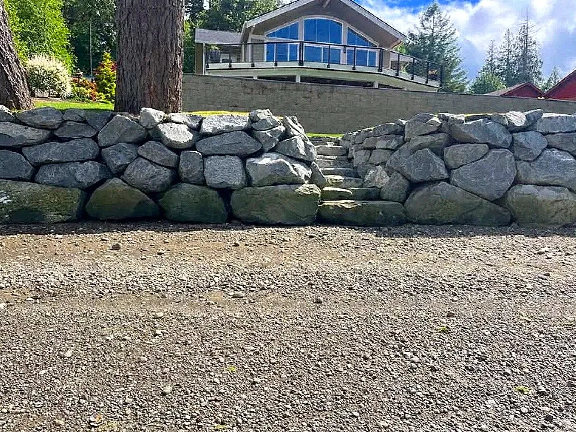 Stone steps leading up to a modern house with large windows and a balcony, surrounded by trees and a gravel area in the foreground.