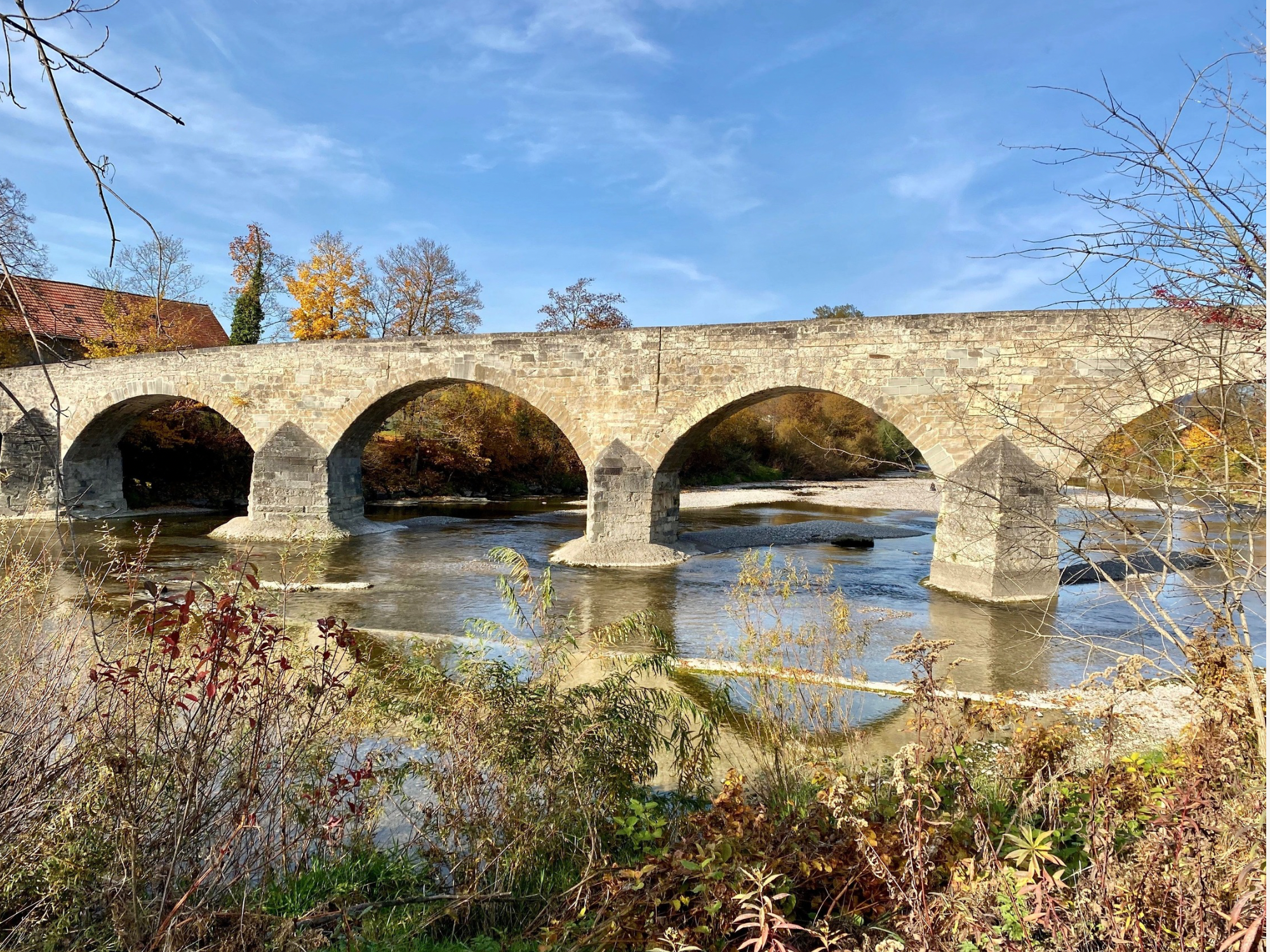 Steinbrücke über einen Fluss mit Bäumen im Herbst, blauer Himmel, einige Wolken.