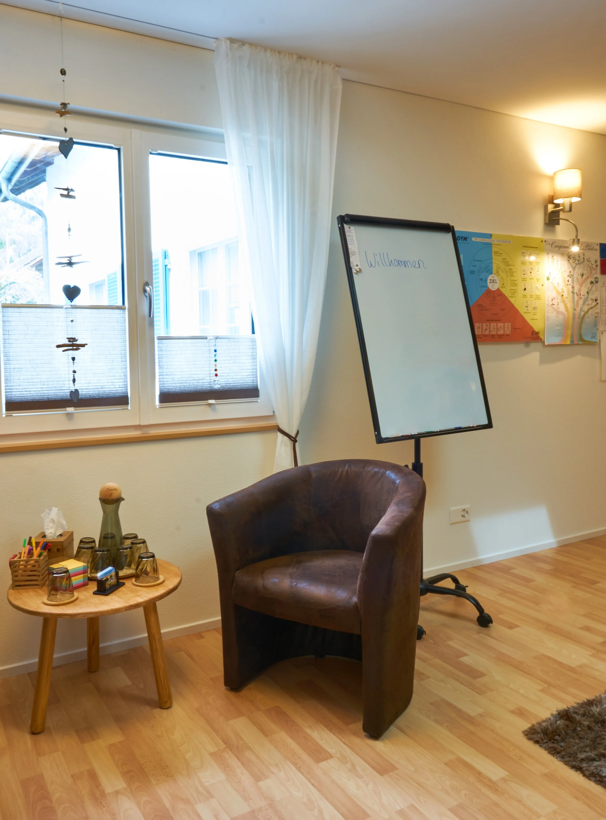 Ein Sitzstuhl, ein kleiner runder Tisch mit Gläser und Schreibutensilien, ein Whiteboard mit der Aufschrift 'Willkommen', sowie Fenster mit Vorhängen und einer Wand mit bunten Postern in einem Raum, vermutlich in einem Seminar- oder Arbeitsbereich.