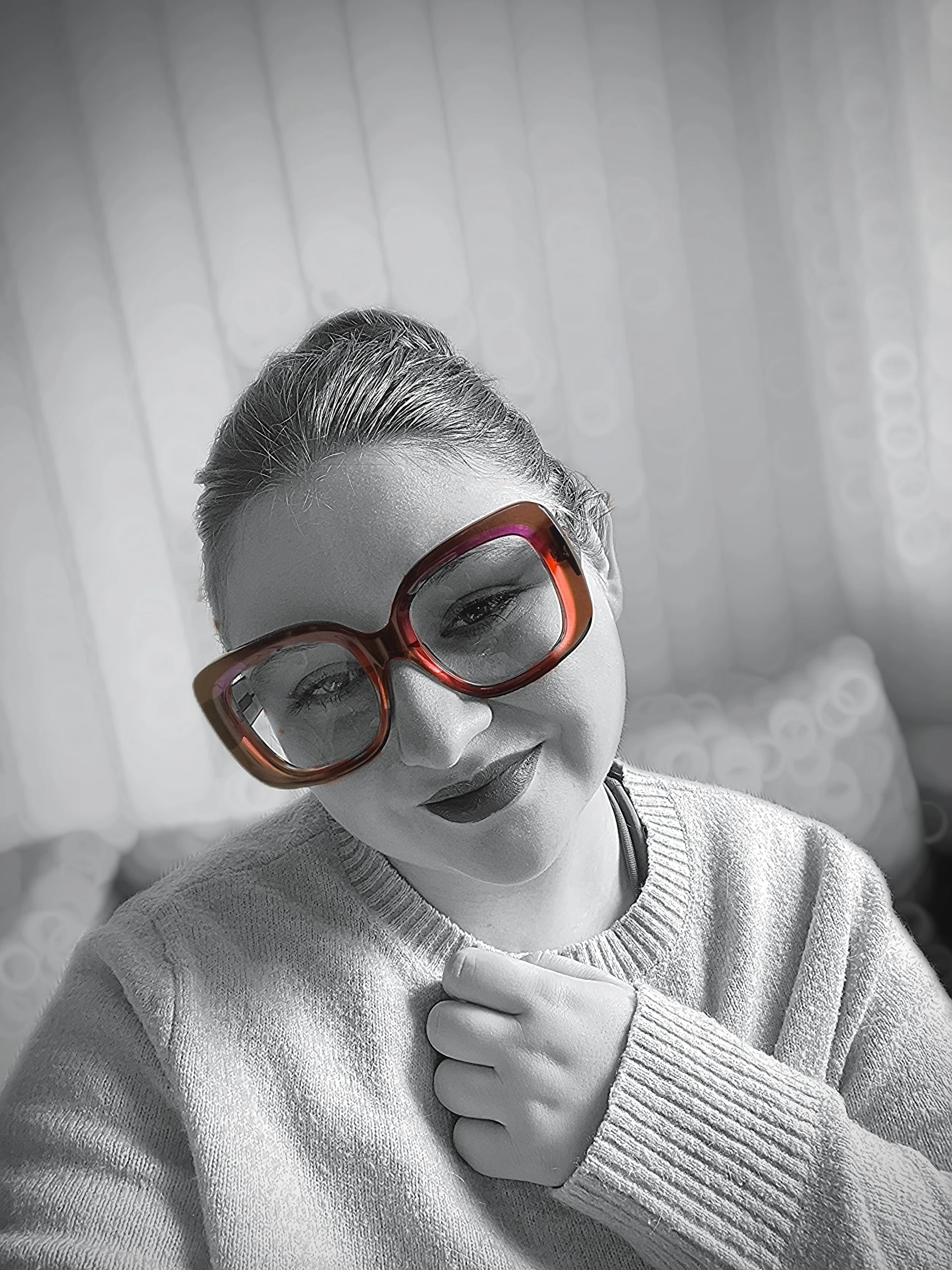 Frau trägt große bunte Sonnenbrille und grau-weißes Strickpullover, lächelt und hält die Hand ans Kinn, Hintergrund unscharf.