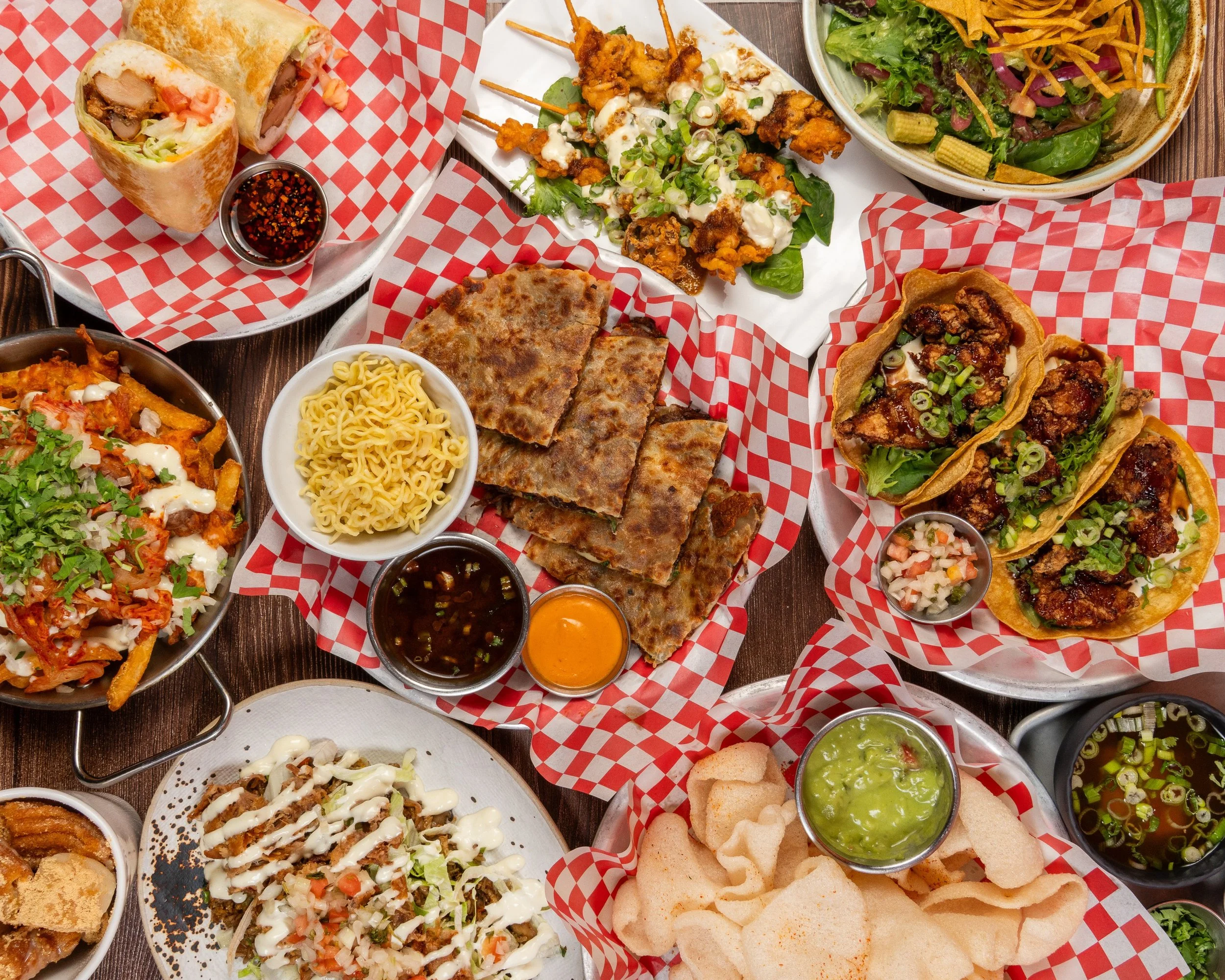 A variety of Mexican dishes including tacos, nachos, burritos, grilled meats, salads, and chips with salsa and guacamole arranged on a wooden table.