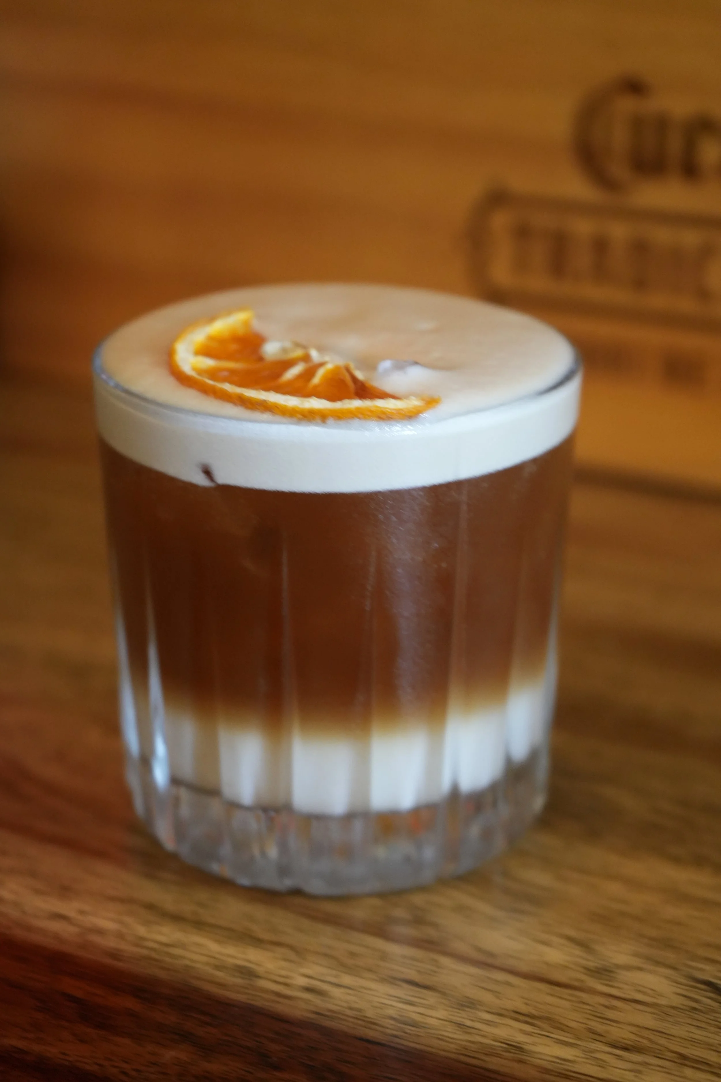 Layered cocktail with whipped cream and orange slice garnish in a short glass on a wooden table.
