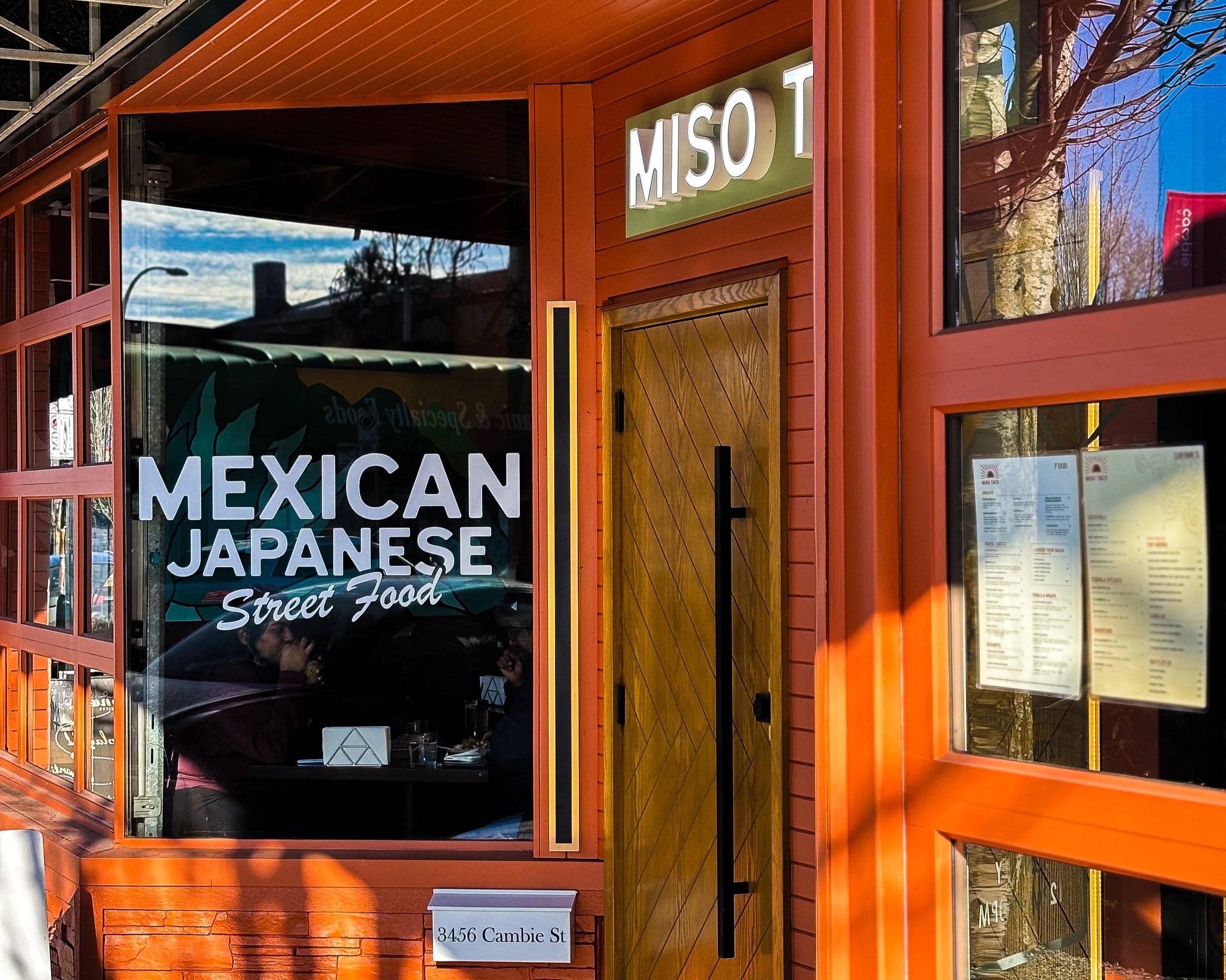 Exterior of a restaurant with orange window frames and walls, signage reading "MISO T" and "Mexican Japanese Street Food", a glass window with silhouettes of people dining inside, and a street number sign "3456 Cambie St".