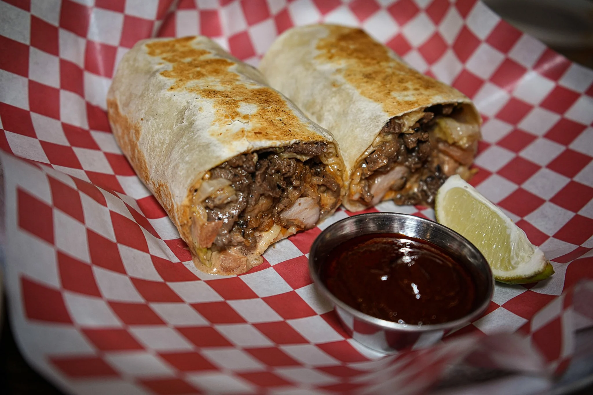 A burrito cut in half showing beef and other ingredients inside, served with a lemon wedge and a small dish of barbecue sauce.