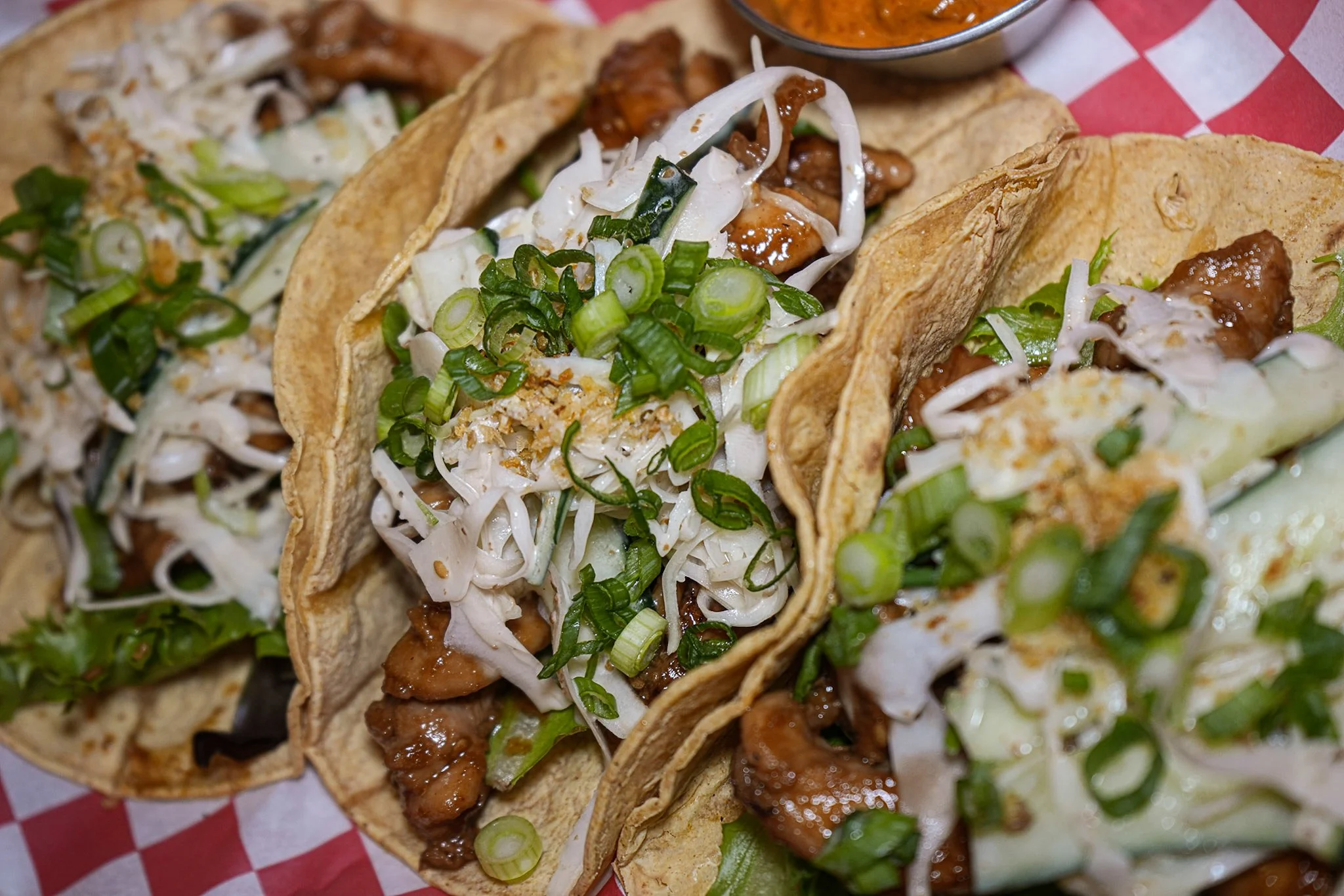 Close-up of three beef tacos on red checkered paper, topped with shredded lettuce and chopped green onions, with a small bowl of salsa in the background.