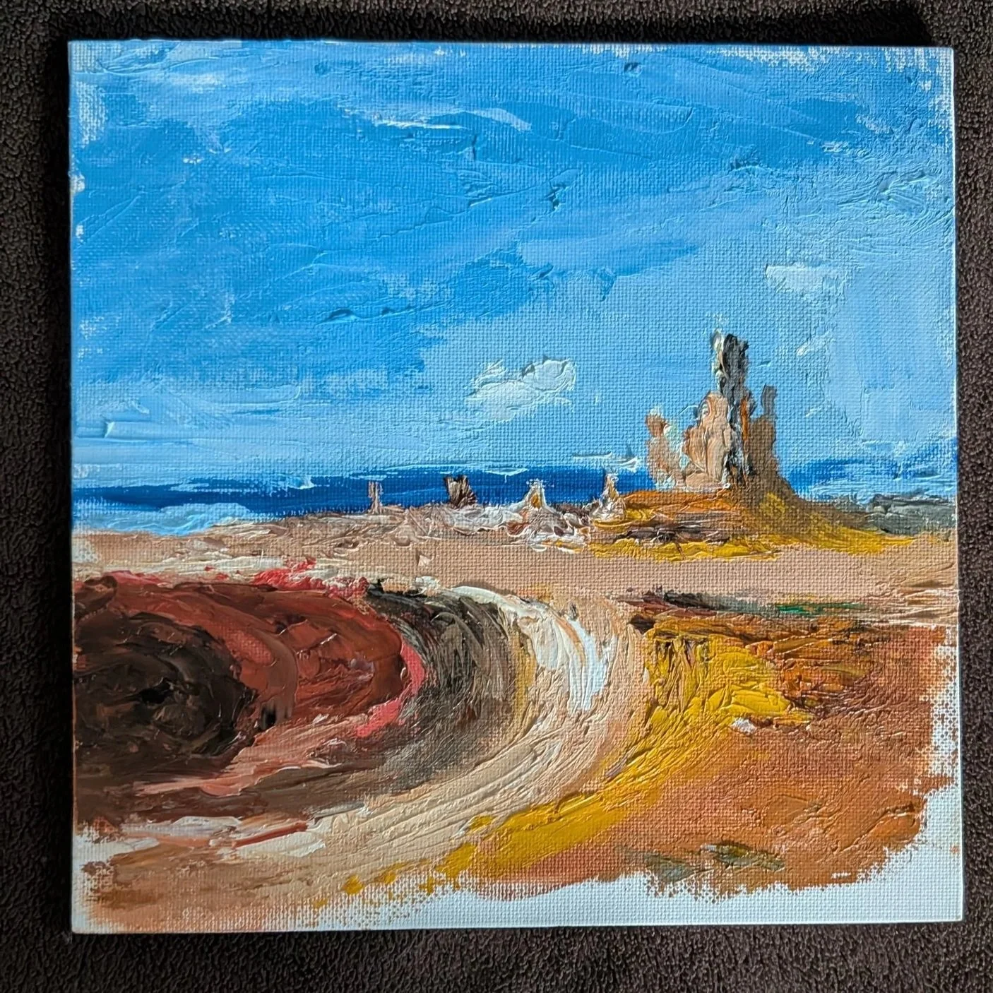 A colorful landscape painting of a beach with a bright blue sky, clouds, a distant ocean, sandy shore, and a large rock formation with smaller rocks around it, painted with bold brushstrokes.