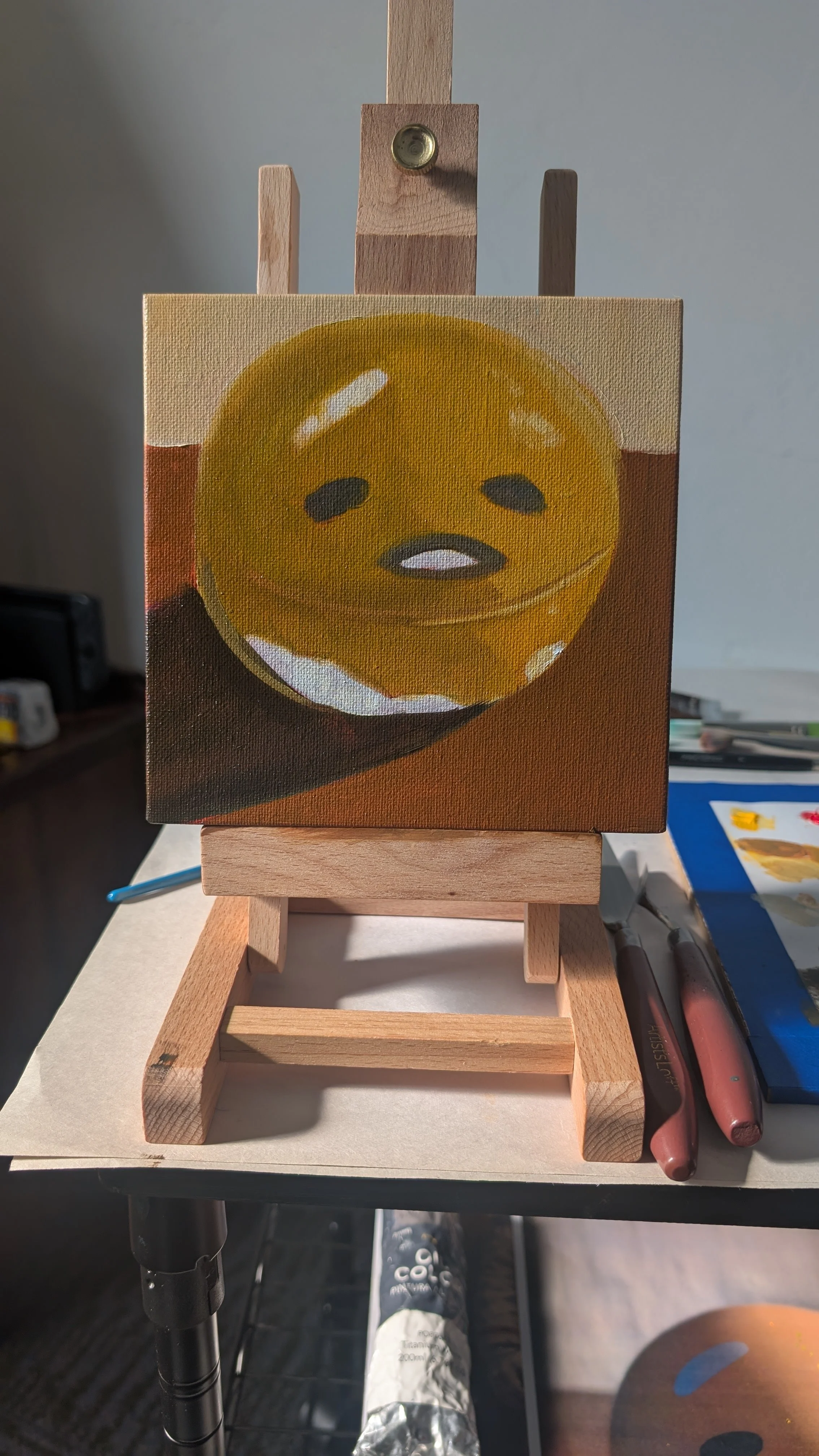 A small painting of a yellow emoji face with closed eyes and a slight frown on a canvas, mounted on a wooden easel.