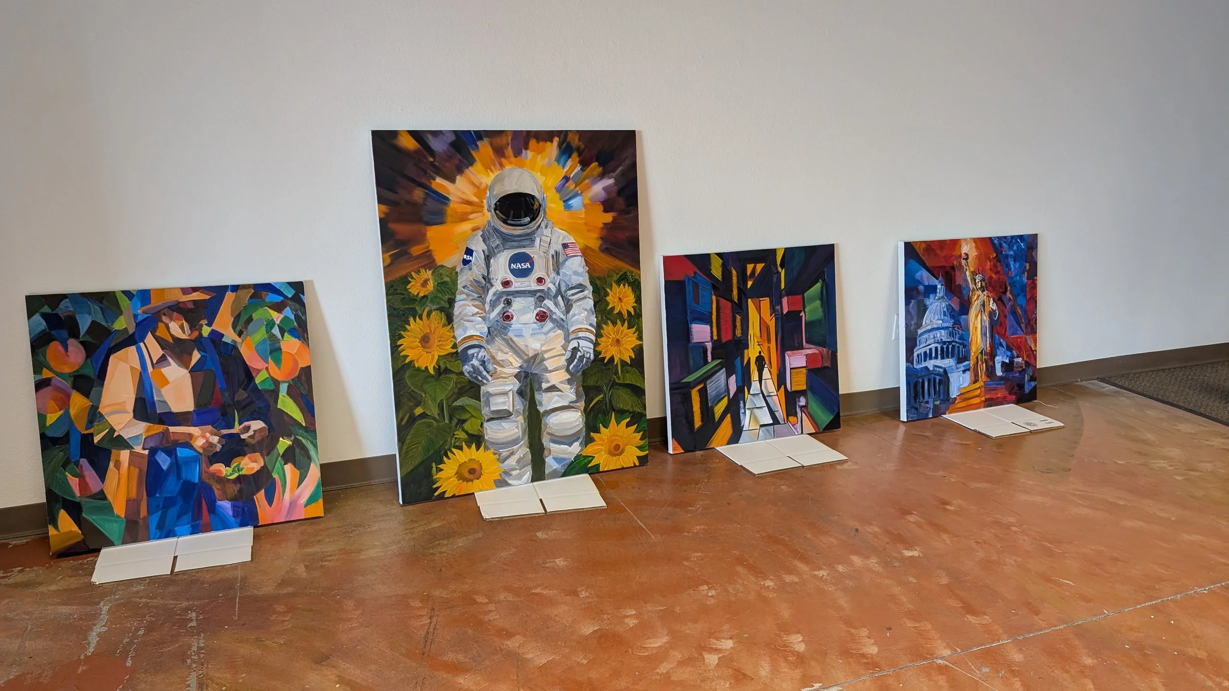 A display of five paintings leaning against a beige wall on a wooden floor. The central painting depicts an astronaut standing among sunflowers with a vibrant, colorful background. The other paintings feature abstract and cityscape scenes.