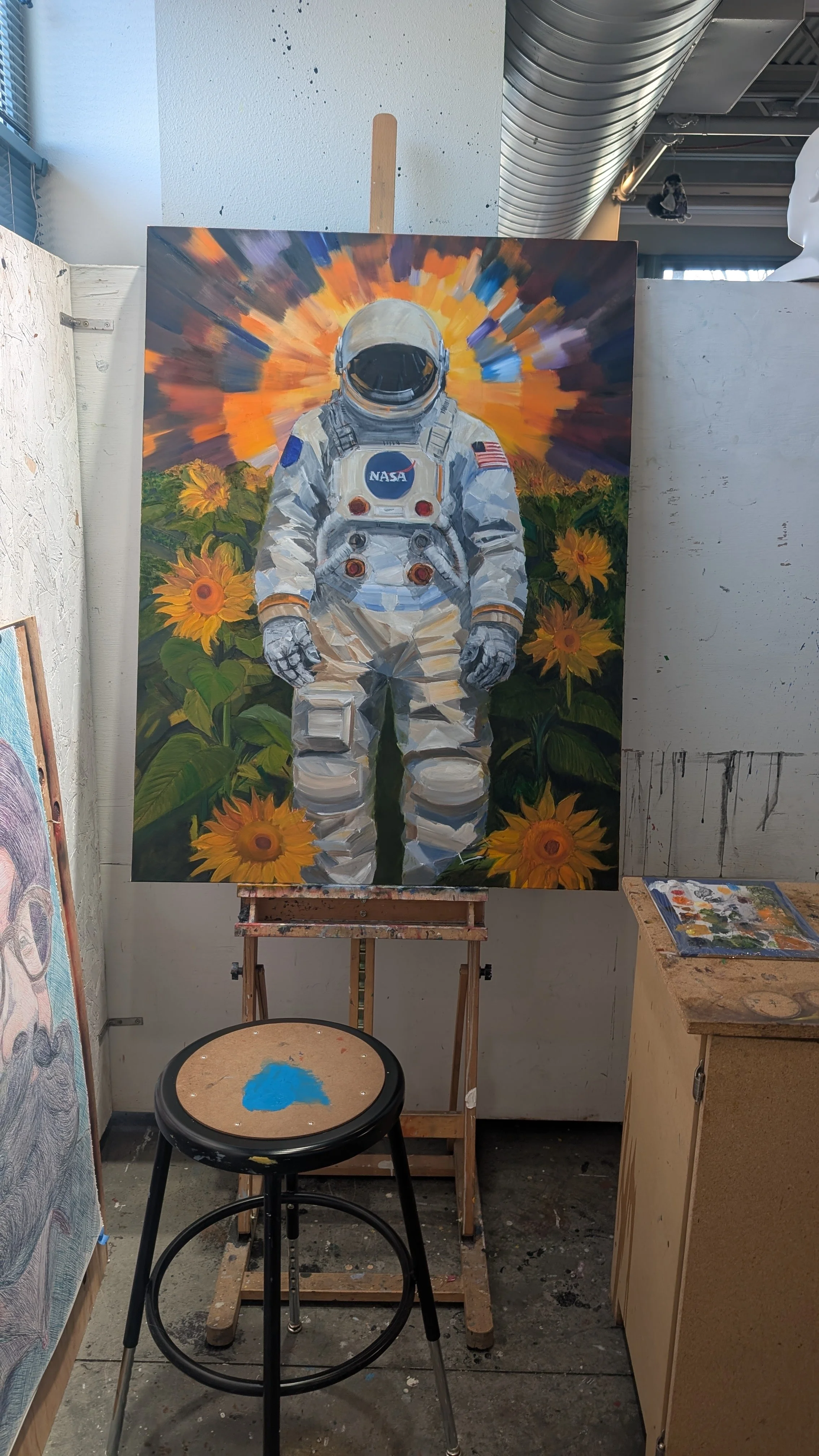 Painting of an astronaut standing in a field of sunflowers with a colorful sky in the background, inside an art studio.