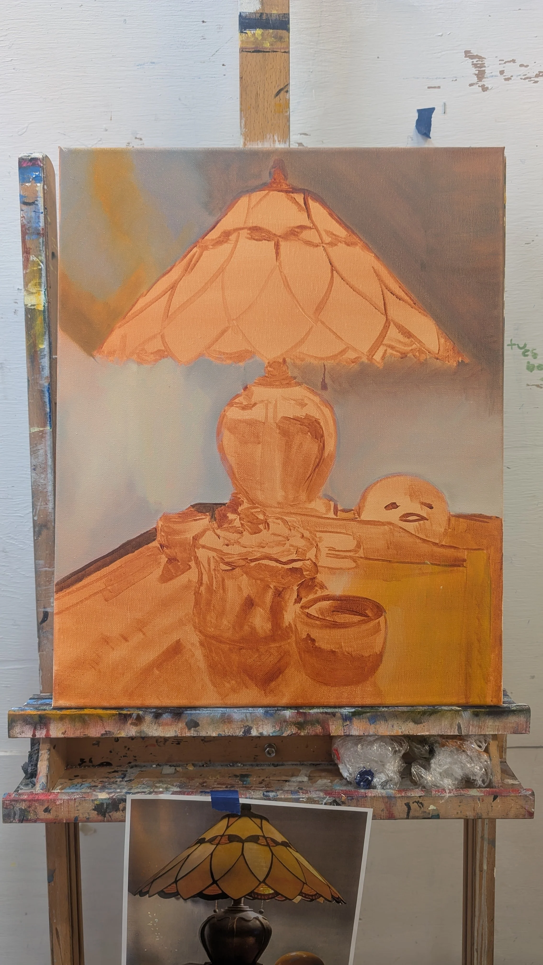 An oil painting of a table lamp on a table with a small cup, a decorative stone, and a ceramic face sculpture. The lamp has a tiled lampshade and is lit, casting a warm glow. The painting is set in an art studio with a wooden easel and a reference ph