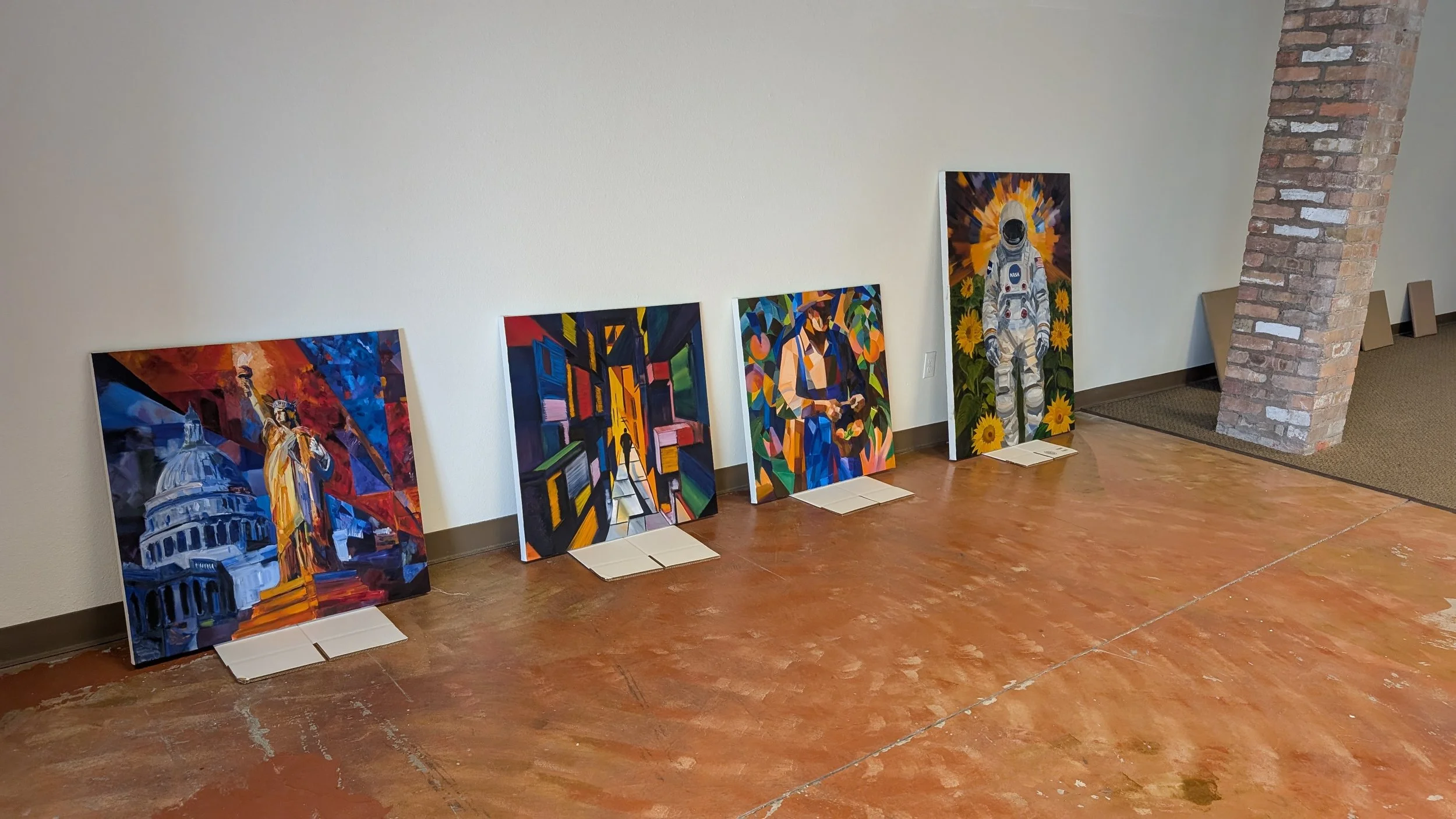 Five colorful paintings are leaning against a plain white wall in a room with a brick corner and wooden floor. All paintings depict scenes related to space or astronauts.