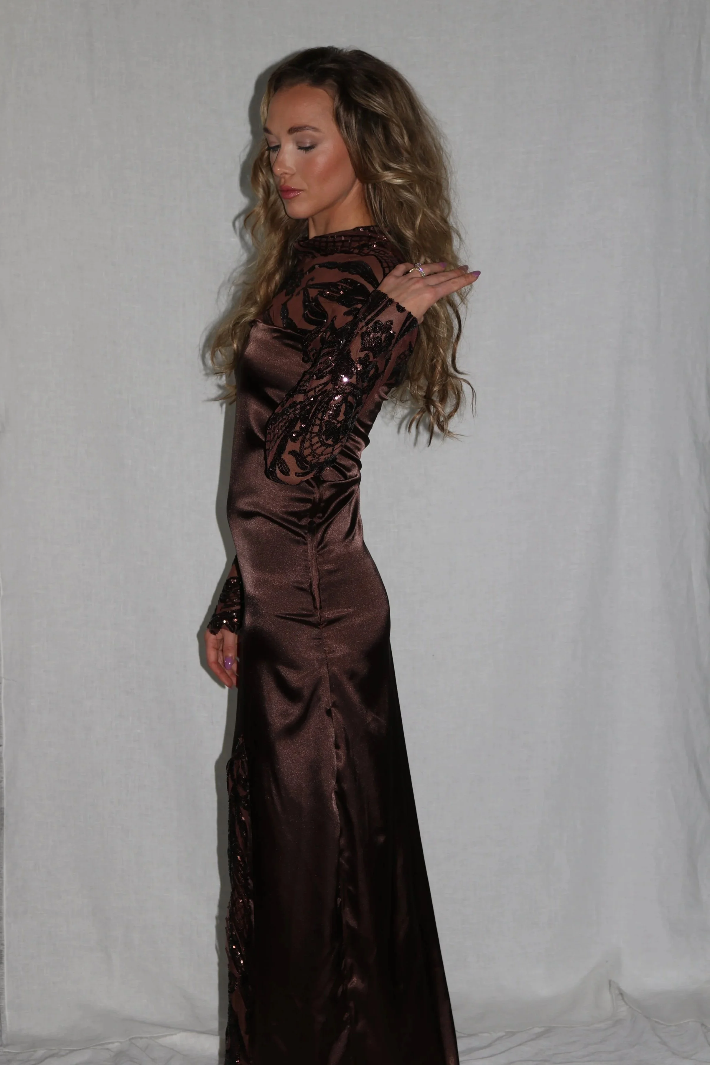 A woman with long, wavy hair wearing a dark satin gown with sheer, embroidered long sleeves, standing against a plain background.
