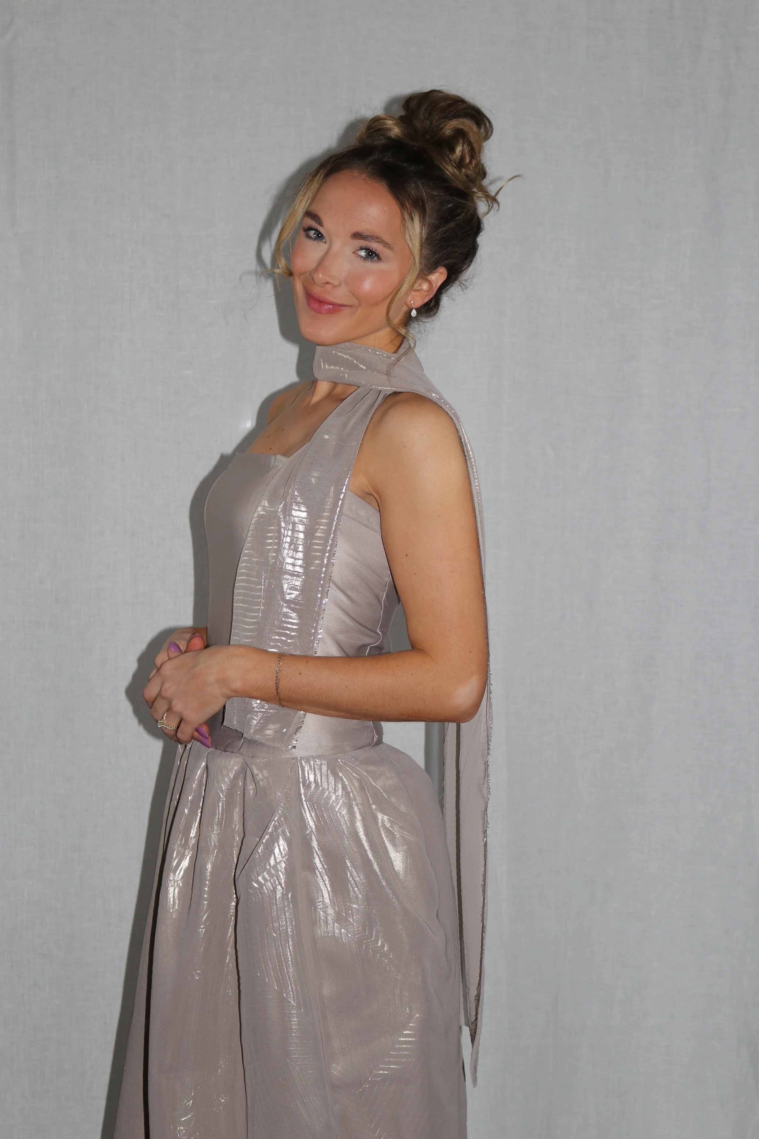 A woman in a shiny, beige sleeveless gown with a matching scarf-like accessory around her neck, smiling and standing against a plain gray background.