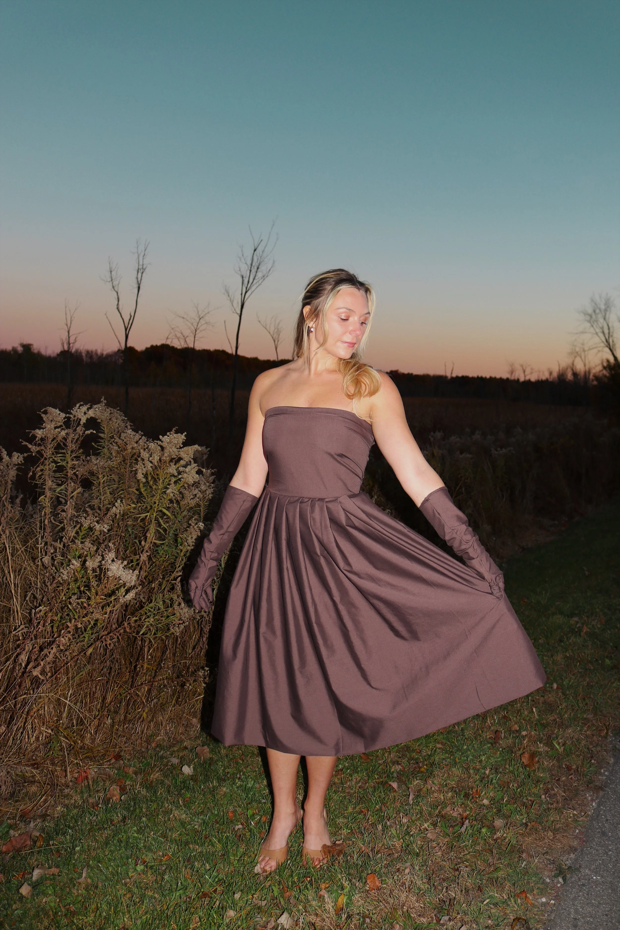 A woman in a strapless brown dress and matching gloves stands outdoors on a grassy area at sunset with a field and leafless trees in the background.