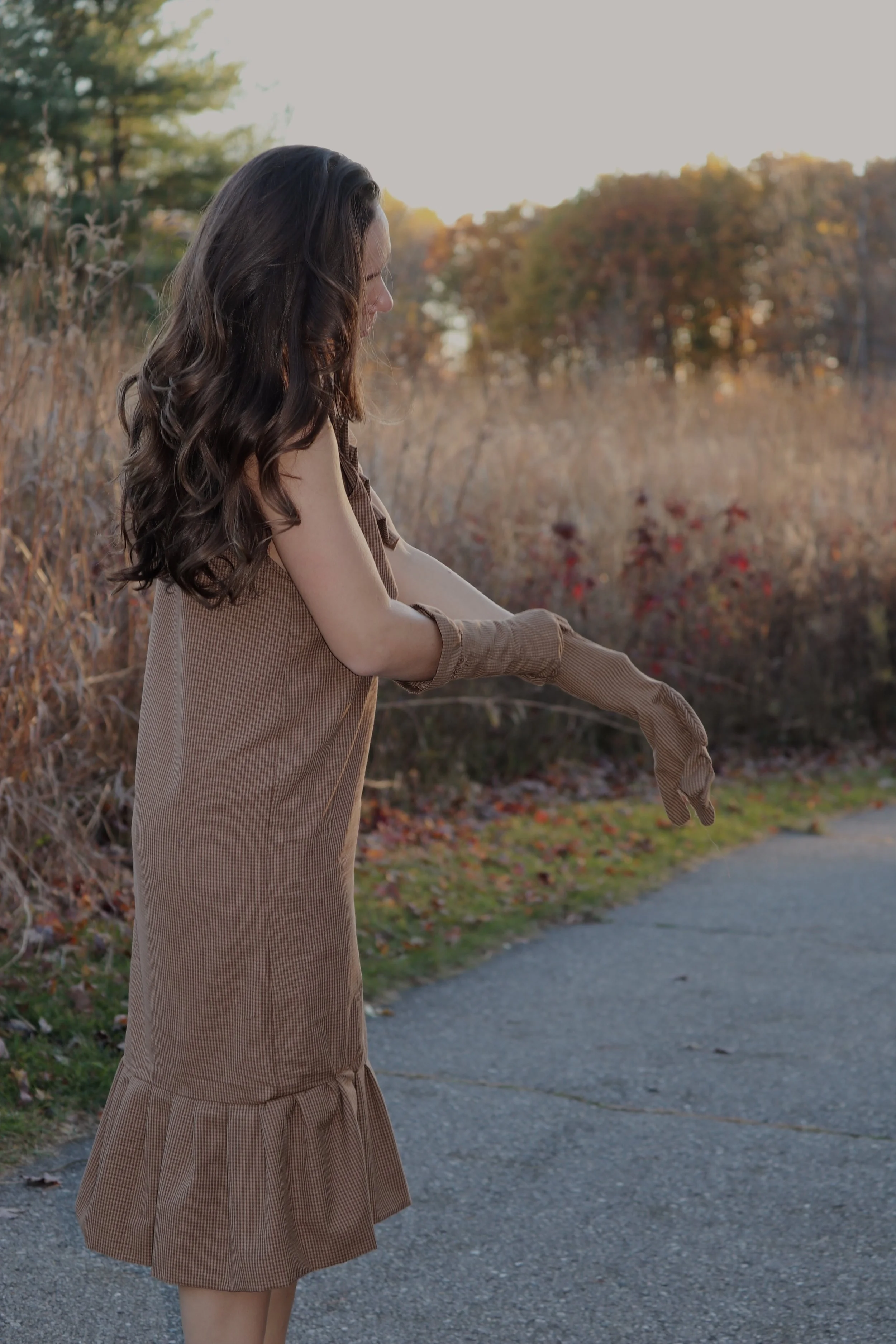 A woman with long, wavy brown hair wearing a vintage brown dress and matching gloves is adjusting her gloves outdoors on a fall day, with trees and dry grass in the background.