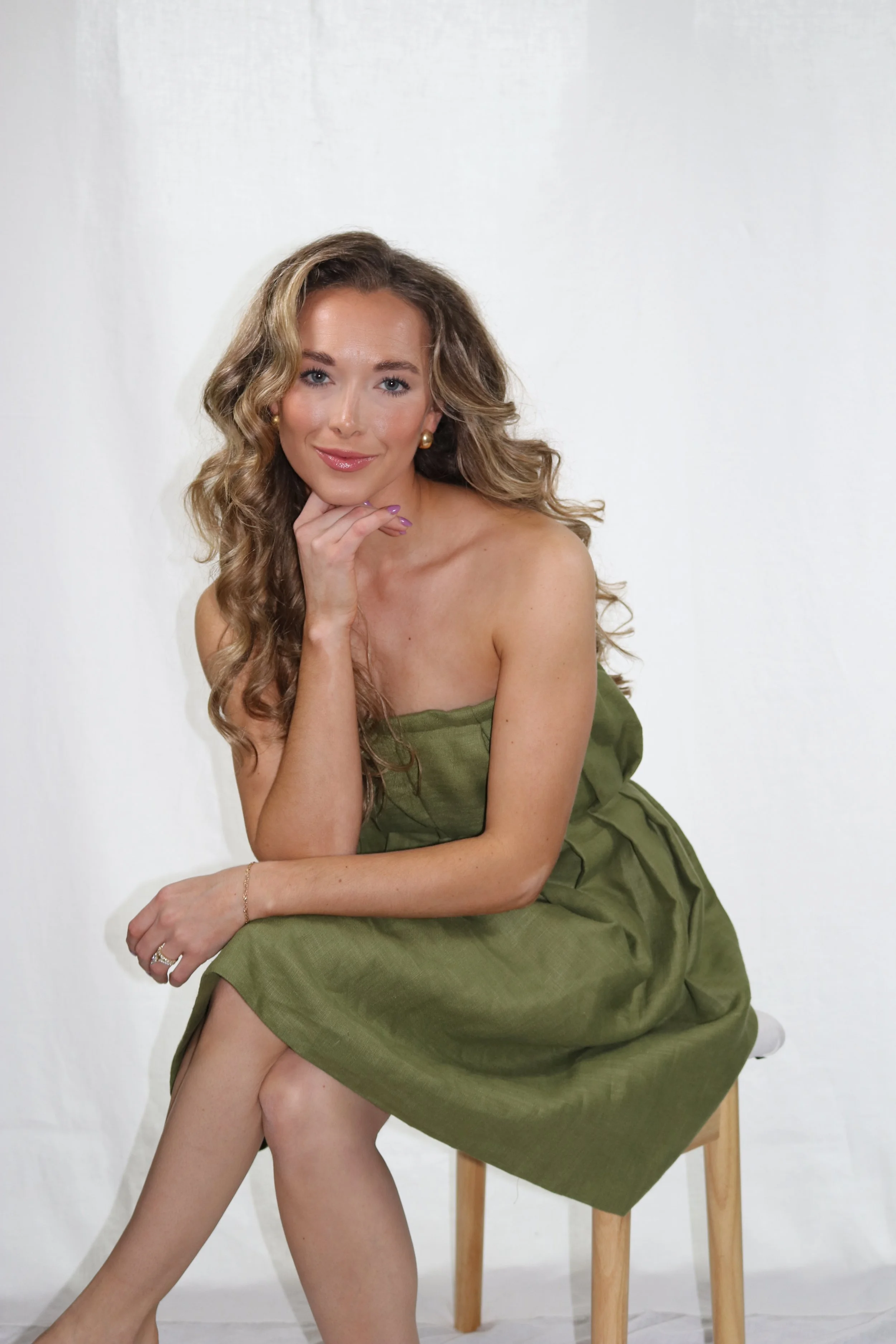 A woman with long, curly blonde hair and blue eyes, sitting on a wooden stool, wearing a green strapless dress, smiling and resting her chin on her hand, against a plain white background.