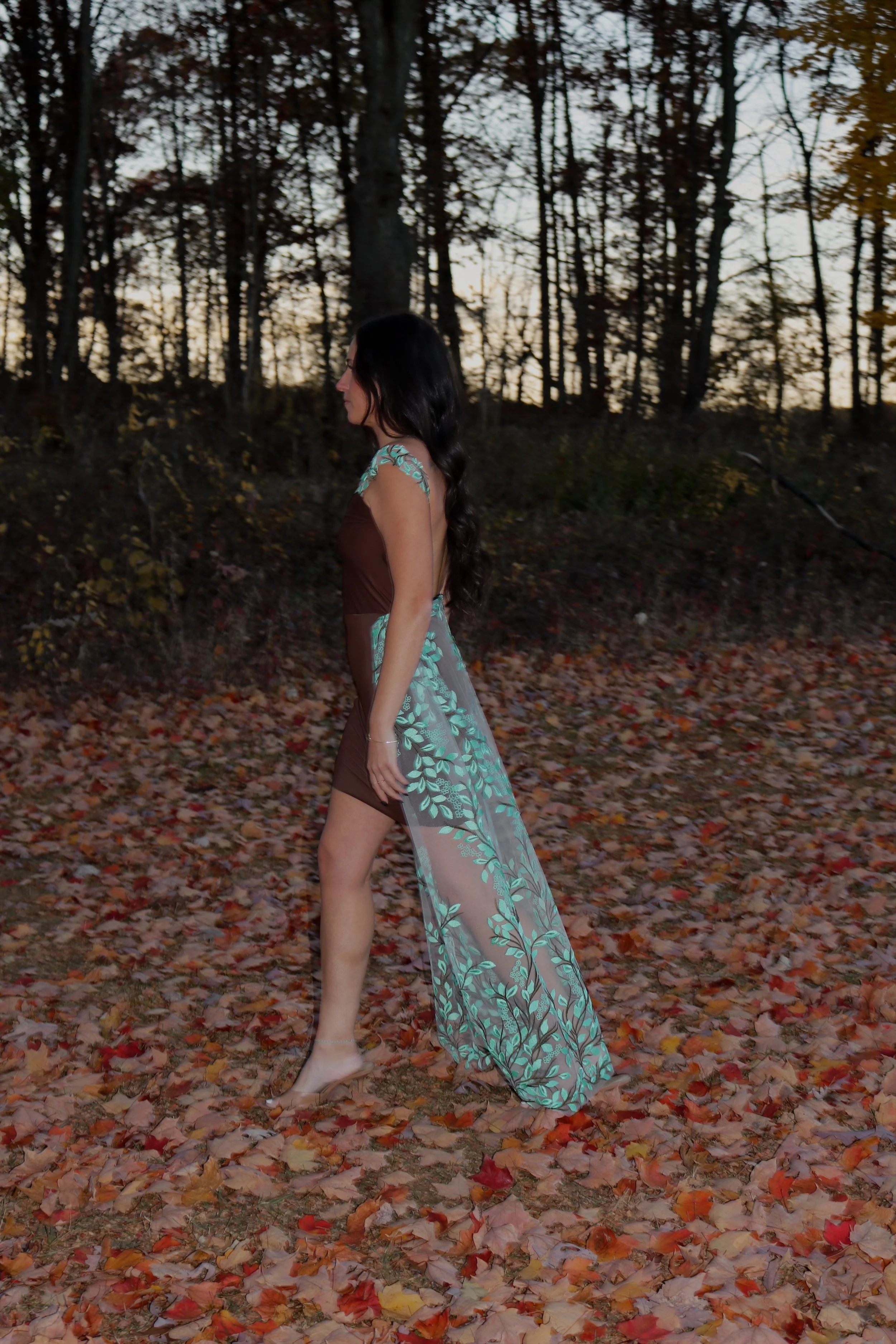 A woman wearing a harness dress with sheer floral detailing walking barefoot on fallen autumn leaves in a wooded area at dusk.