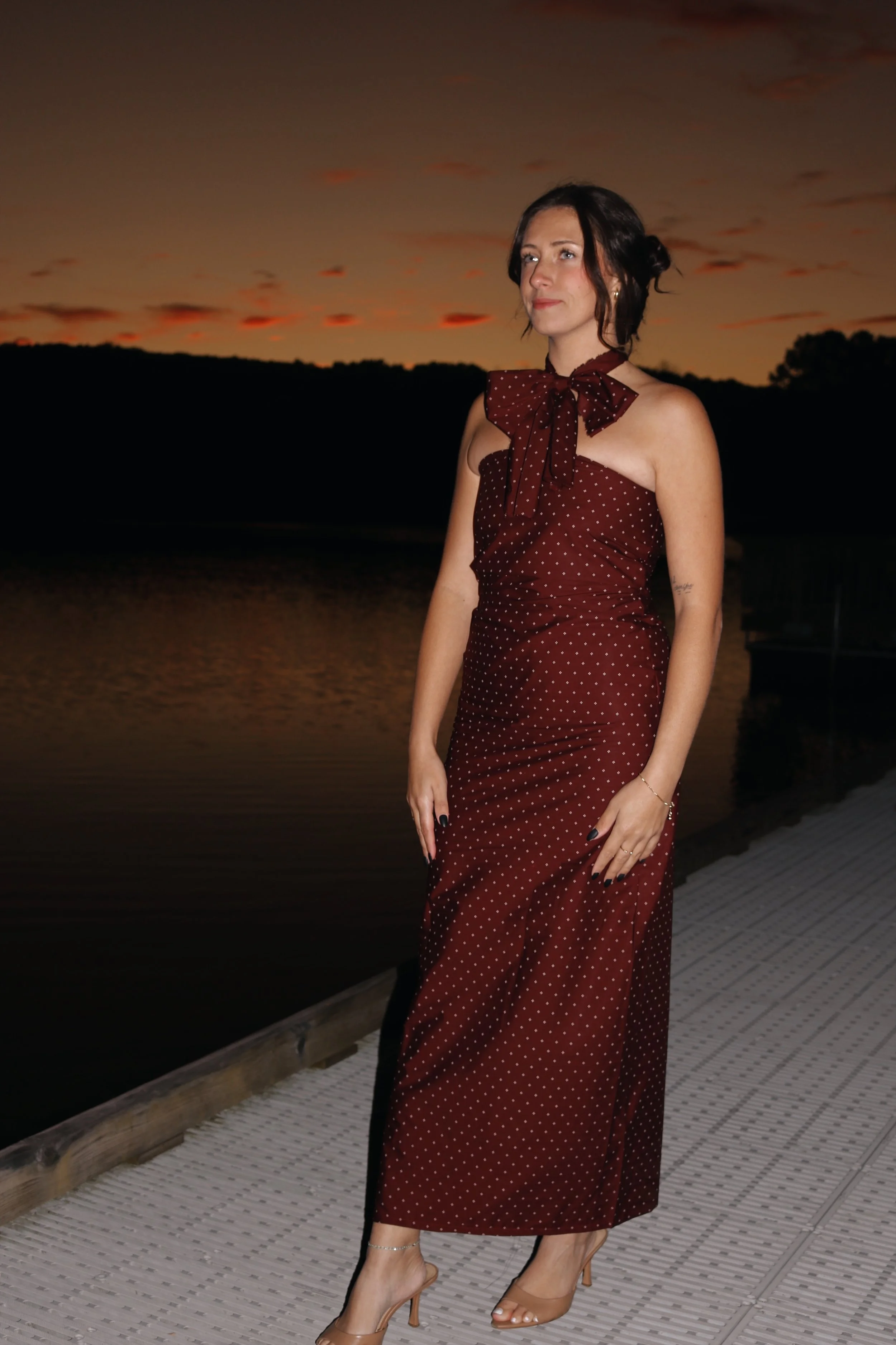 A woman in a burgundy polka dot dress standing on a dock at sunset, near a body of water.