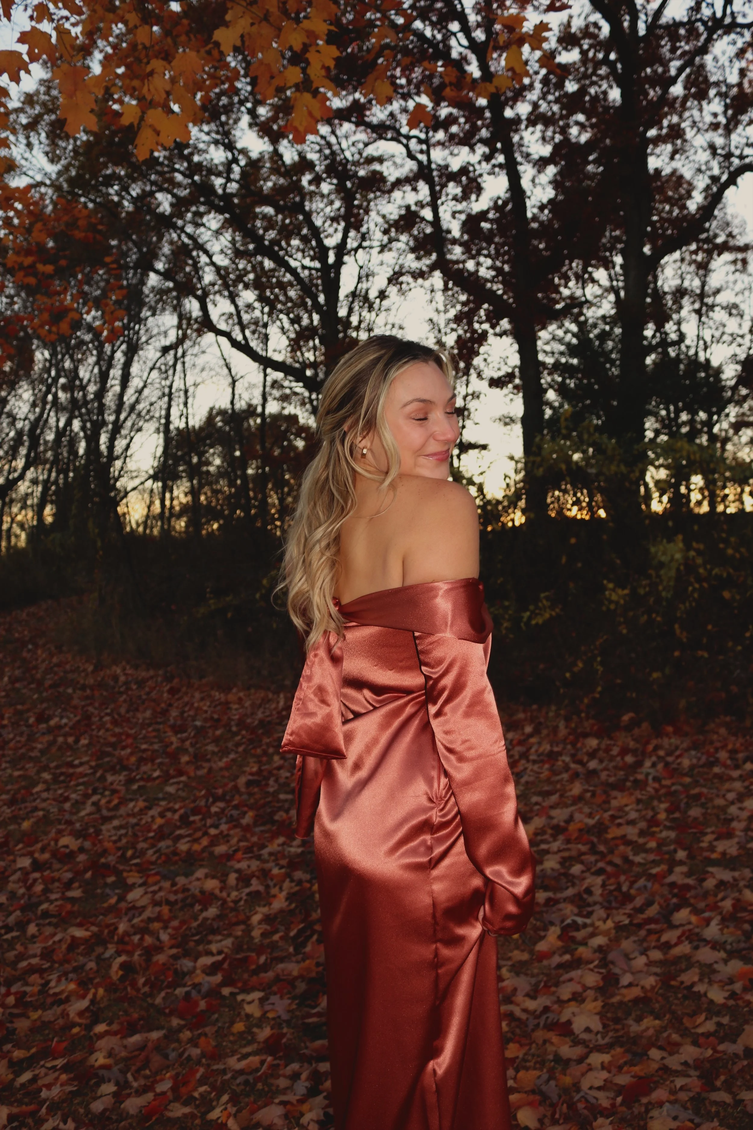 A woman in a satin, off-shoulder, rust-colored dress standing outdoors on a fall evening, surrounded by trees with orange and yellow leaves, with a sunset in the background.