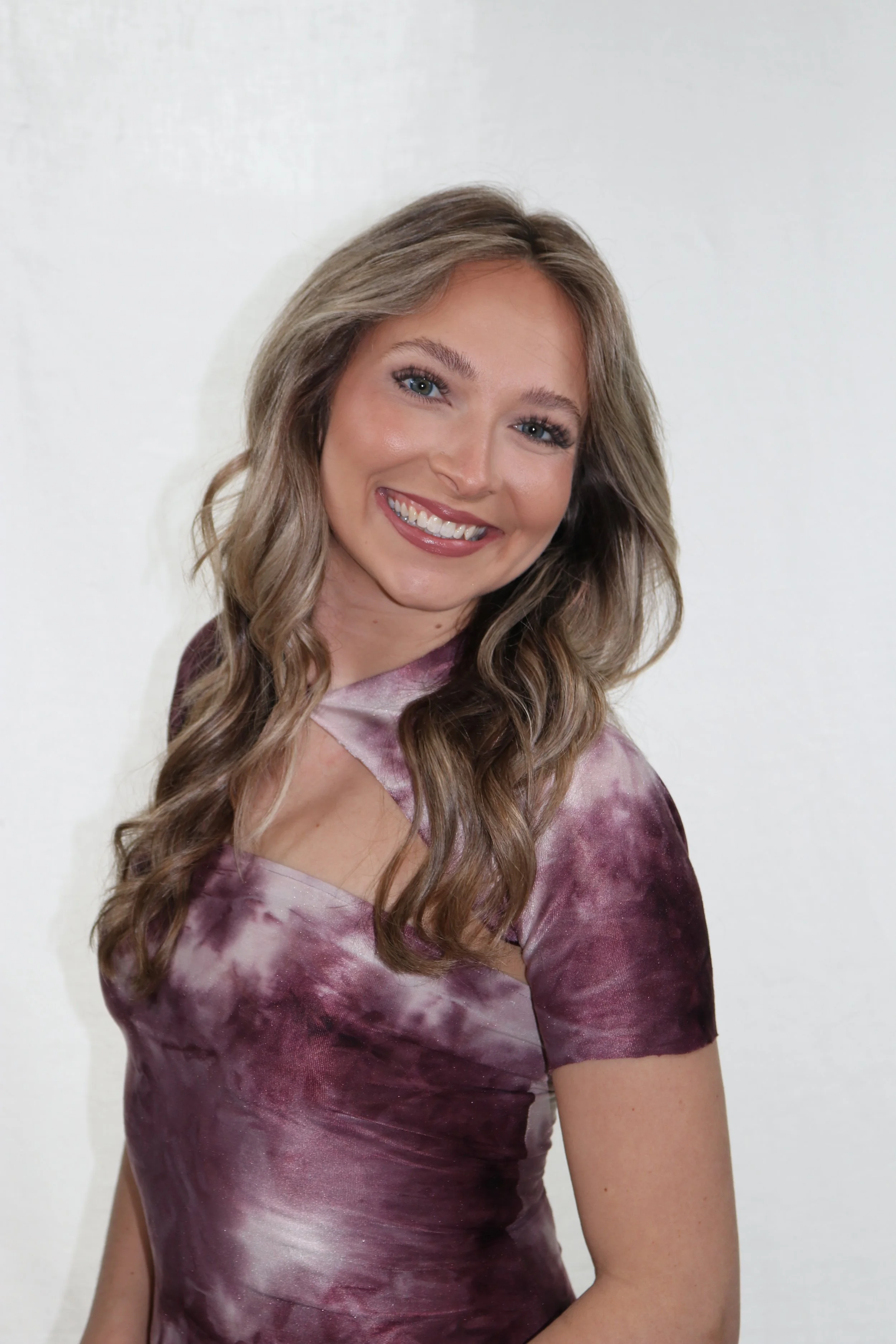 A woman with wavy, shoulder-length blonde hair smiling at the camera, wearing a maroon and pink tie-dye dress, standing against a plain white background.
