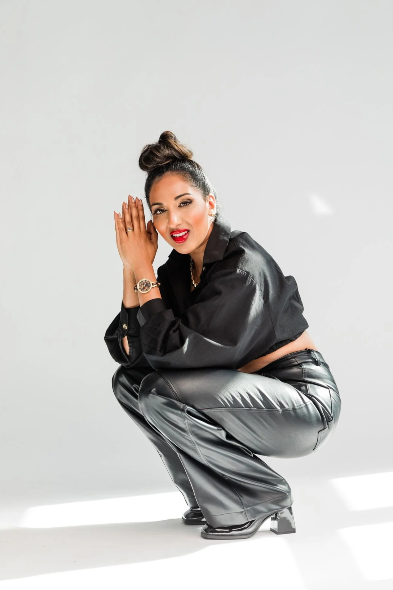 A woman squatting in a crouch, wearing a black satin shirt, black leather pants, and high-heeled shoes, with her hands near her face, smiling and looking at the camera, against a plain white background.