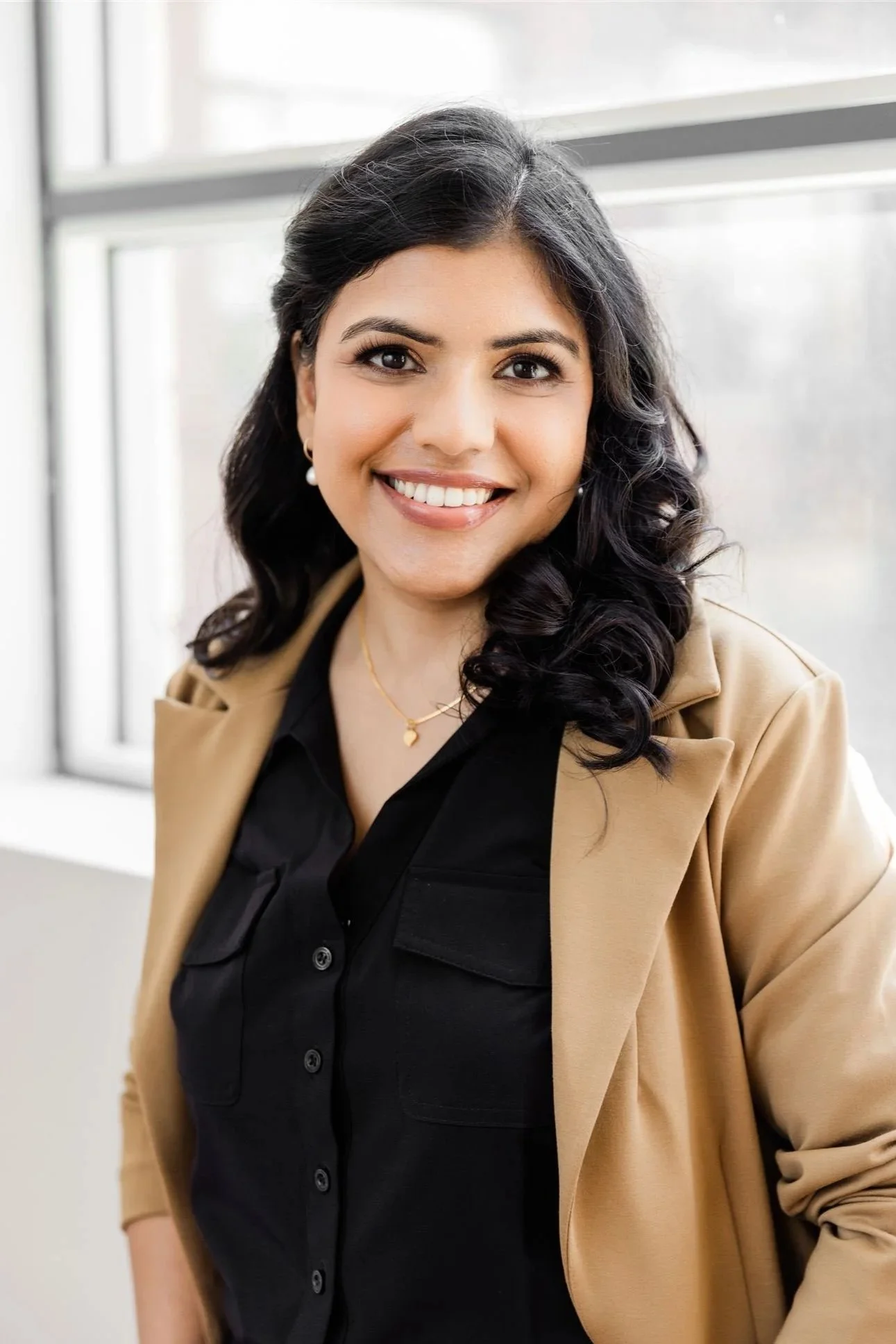 A woman with long black hair and light skin, smiling, wearing a black shirt and tan blazer, standing indoors near a window.