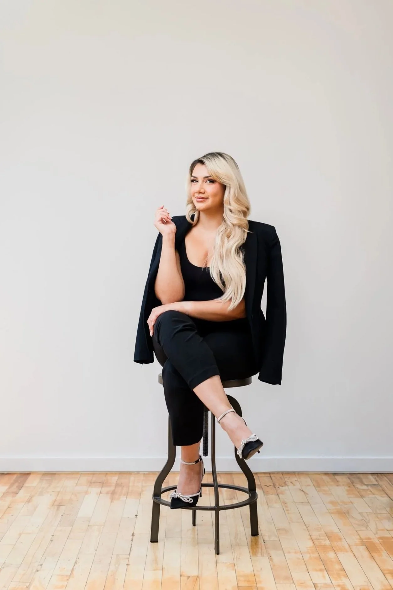 A woman with long blonde hair, wearing a black outfit and heels, sitting on a stool with one leg crossed over the other, posing against a plain white wall.