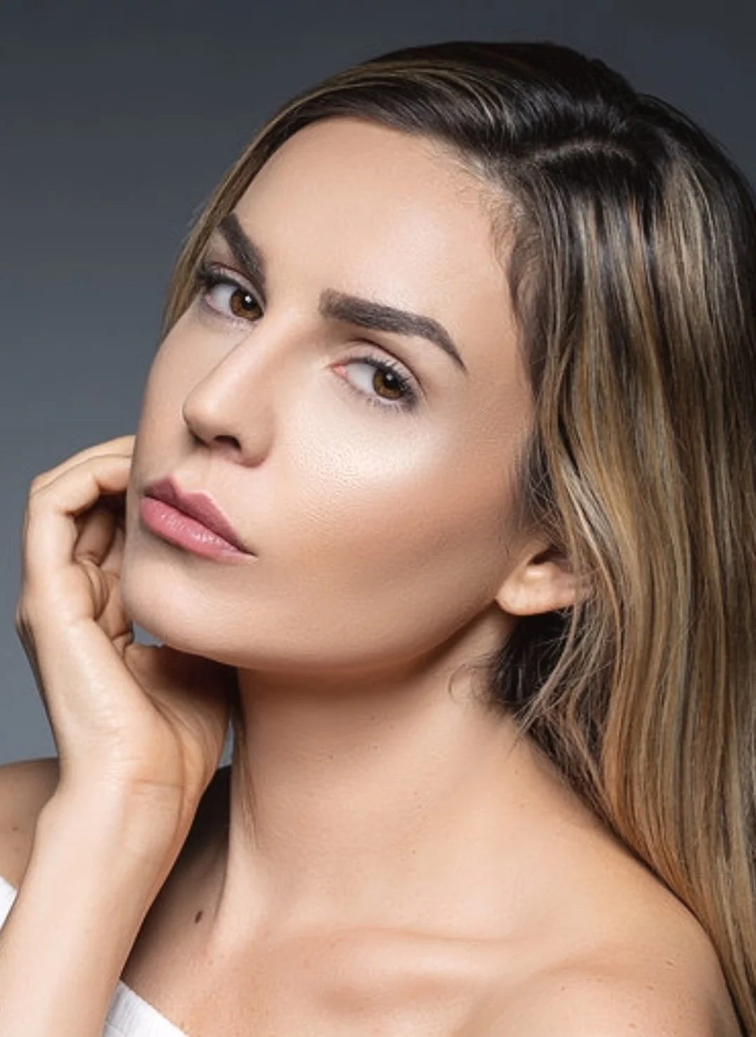 Close-up portrait of a woman with light skin, brown eyes, and brown hair with highlights, posing with her hand near her face against a gray background.